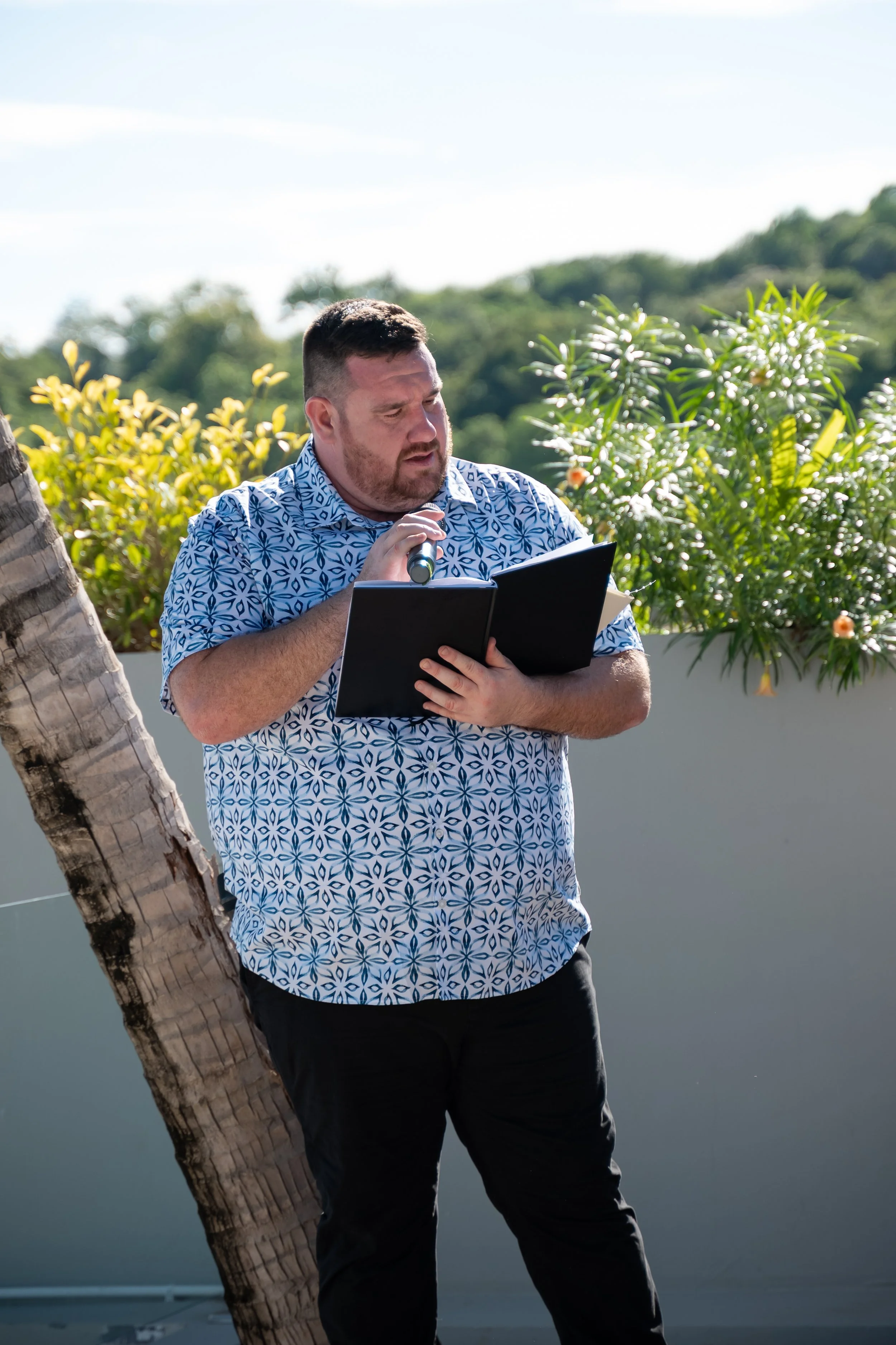 A man in a blue patterned shirt is holding a microphone and reading from a black notebook outdoors. There are lush green trees and plants in the background.
