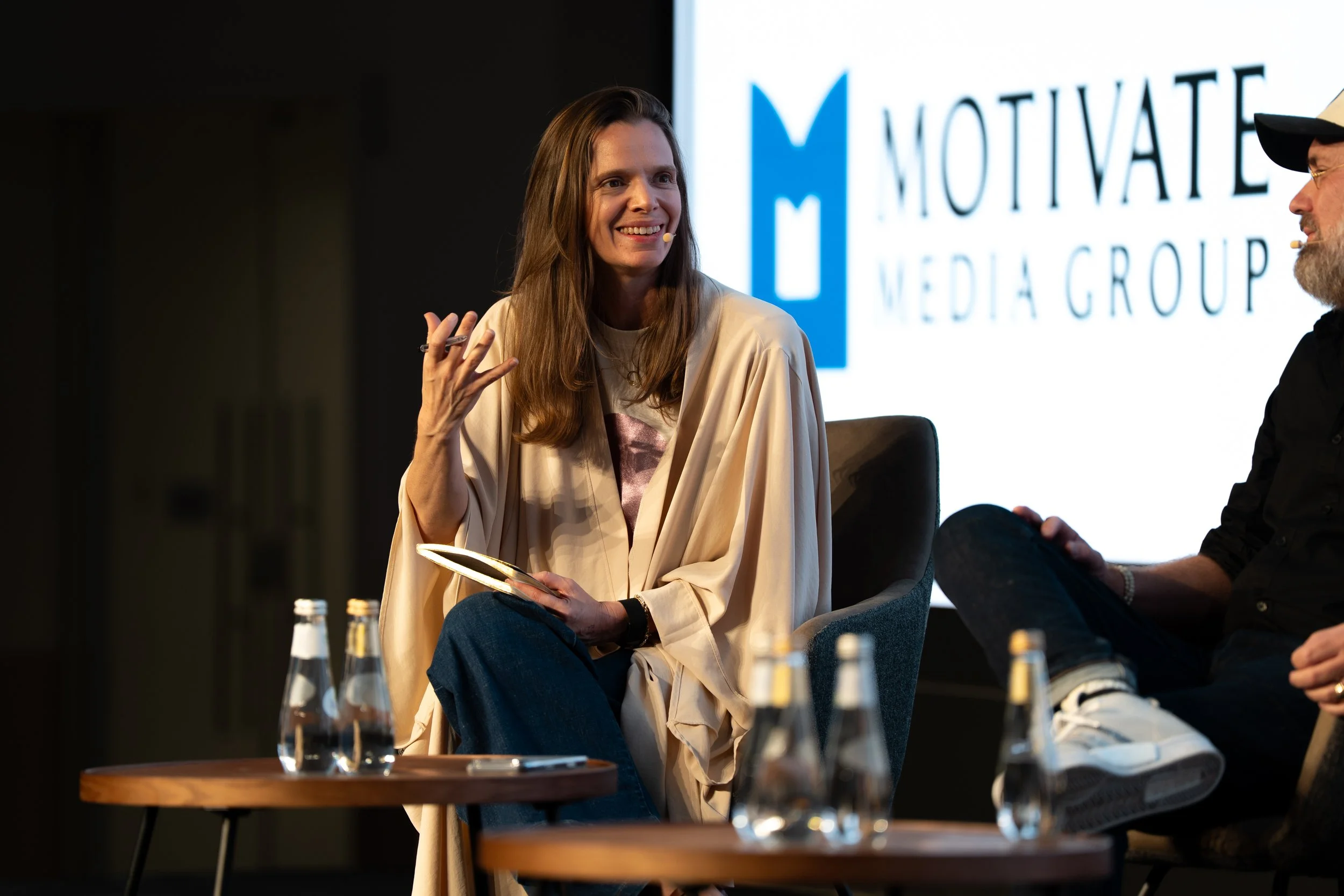 A woman (Lucy Harvey) with long brown hair smiling, sitting moderating a panel during a presentation or discussion, with a man wearing glasses and a cap sitting beside her.