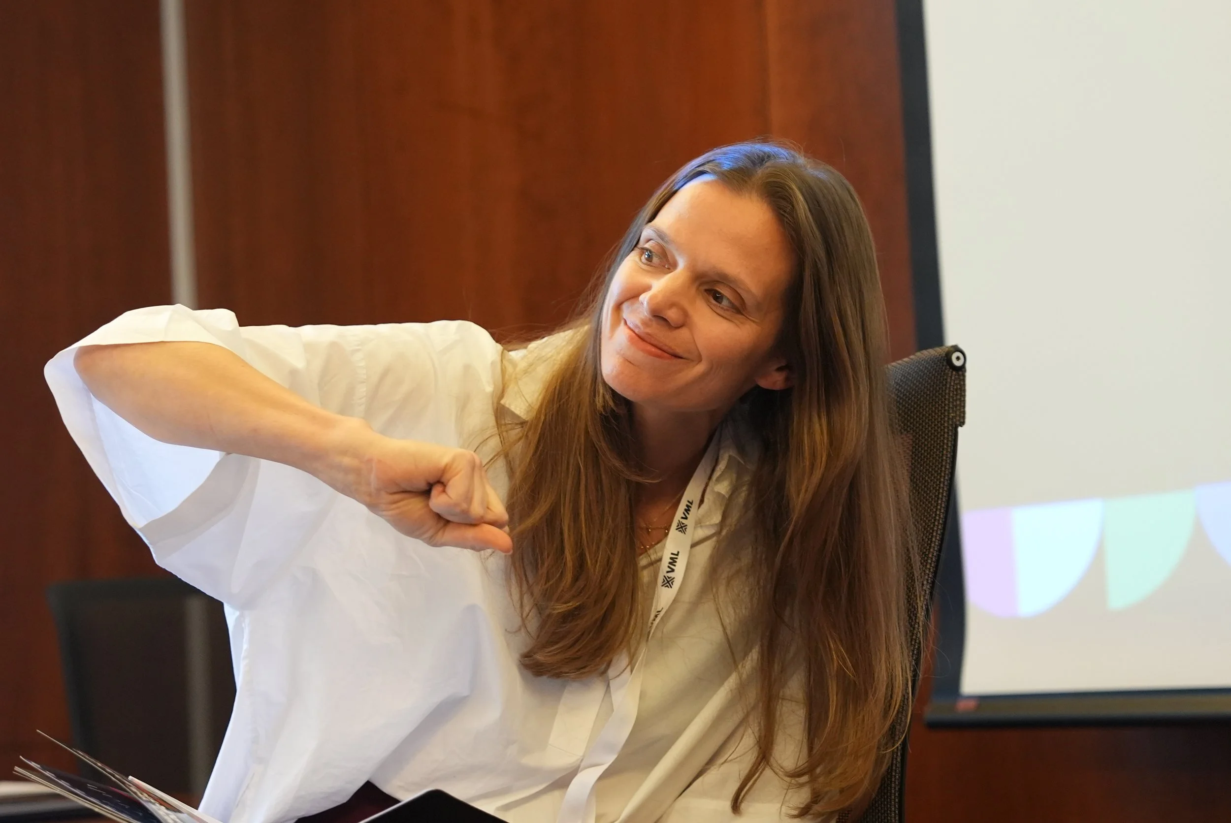 A woman (Lucy Harvey) with long brown hair is smiling and giving a fist bump whilst facilitating a meeting or workshop. Wearing a white shirt and a lanyard, seated in a conference room.