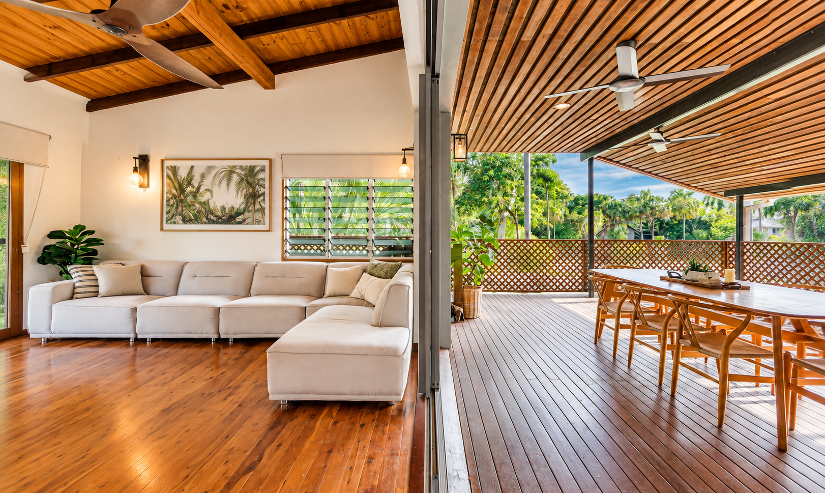 The image shows an open-concept living room and outdoor patio separated by a glass sliding door. The living room has a white sectional sofa, wood flooring, a framed tropical landscape picture on the wall, and indoor plants. The patio has wooden flooring, a large wooden dining table with chairs, and a lattice railing. Both areas feature wooden ceilings with ceiling fans, and lush greenery is visible outside.
