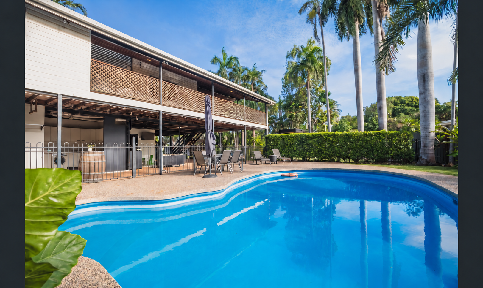 A backyard with a swimming pool, surrounded by greenery and tall palm trees. There is an elevated house with a patio, outdoor dining area, and lounge chairs.
