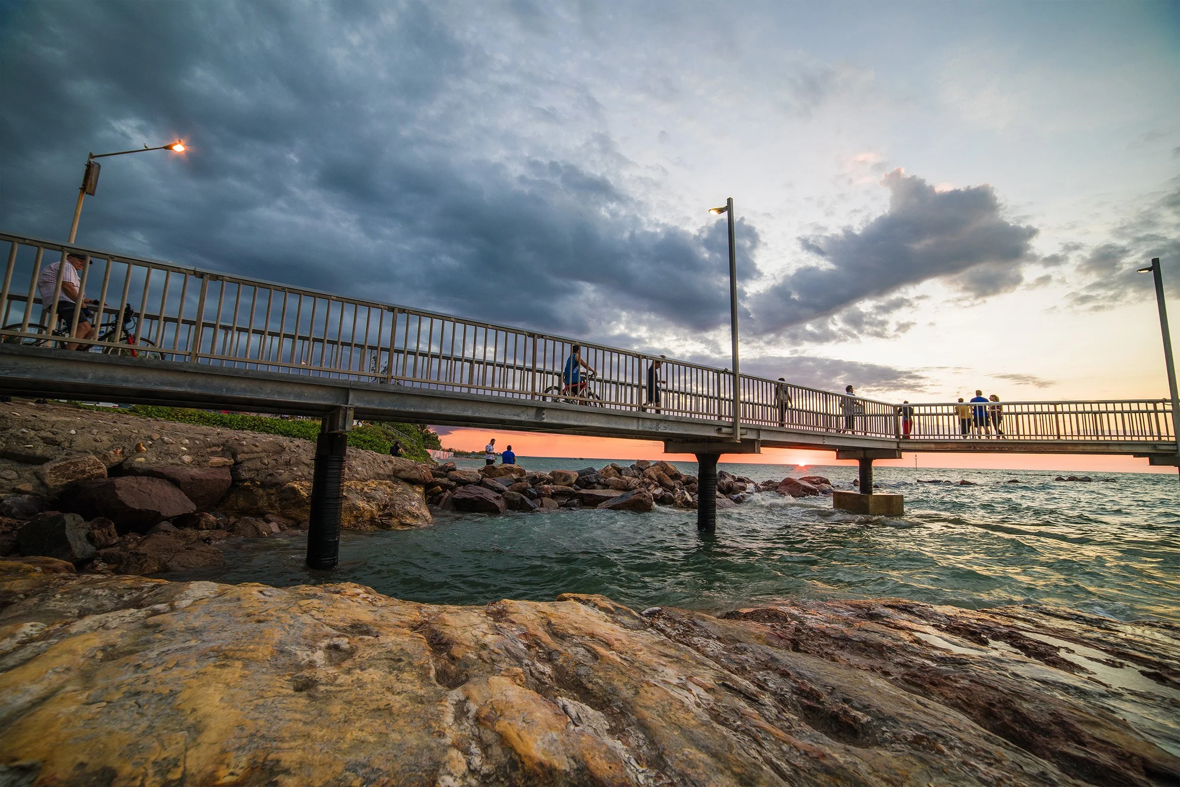 People walking and riding bicycles on a bridge over rocky shore during sunset, with dark clouds and a partly cloudy sky.