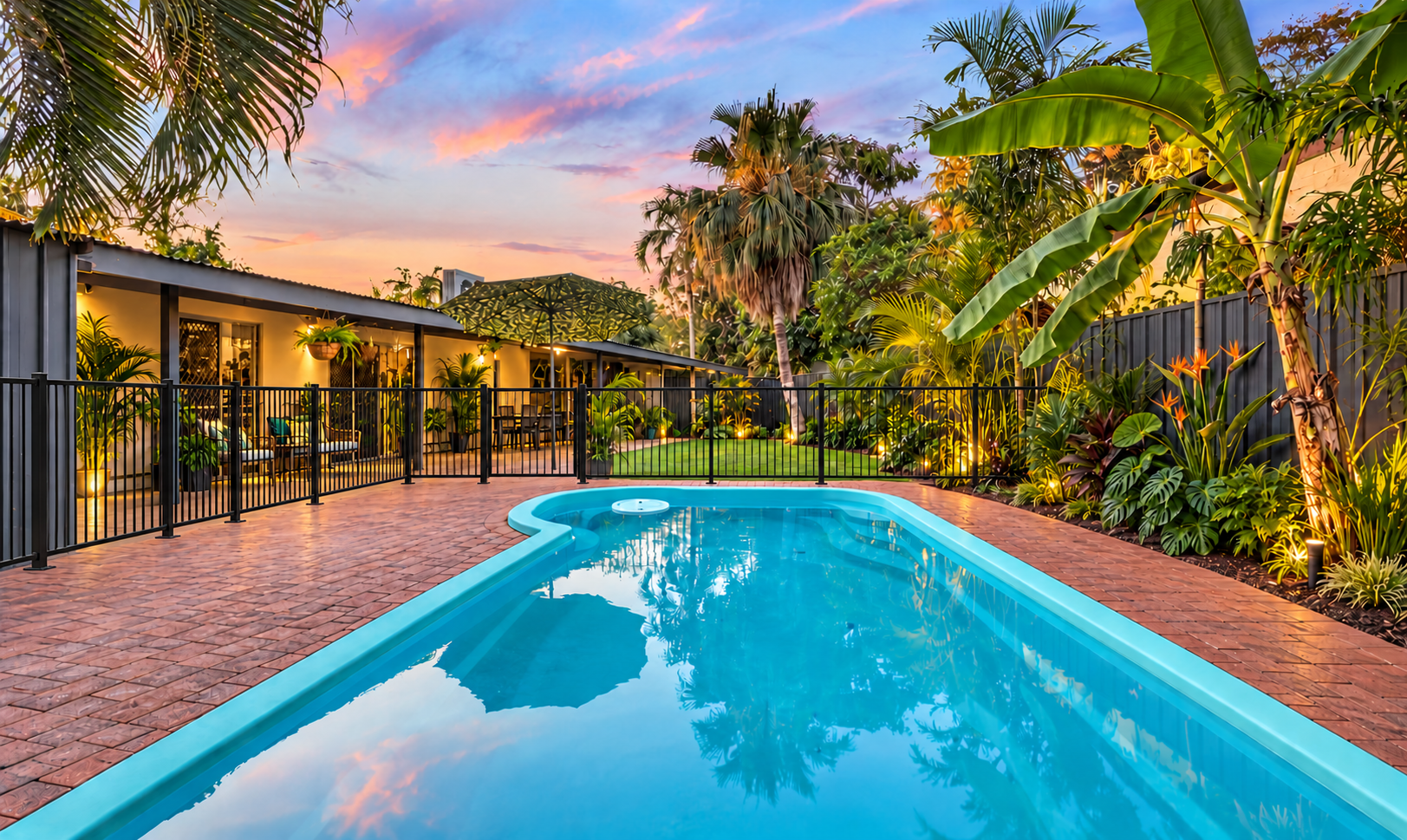 A backyard with a swimming pool, surrounded by lush tropical plants and trees, with patio furniture and a large umbrella, during sunset with colorful sky.