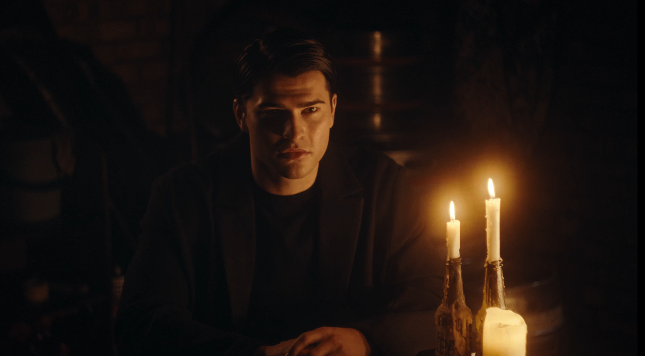 A young man with dark hair sitting in dim candlelight, looking at the camera with a serious expression, next to a table with three lit candles.