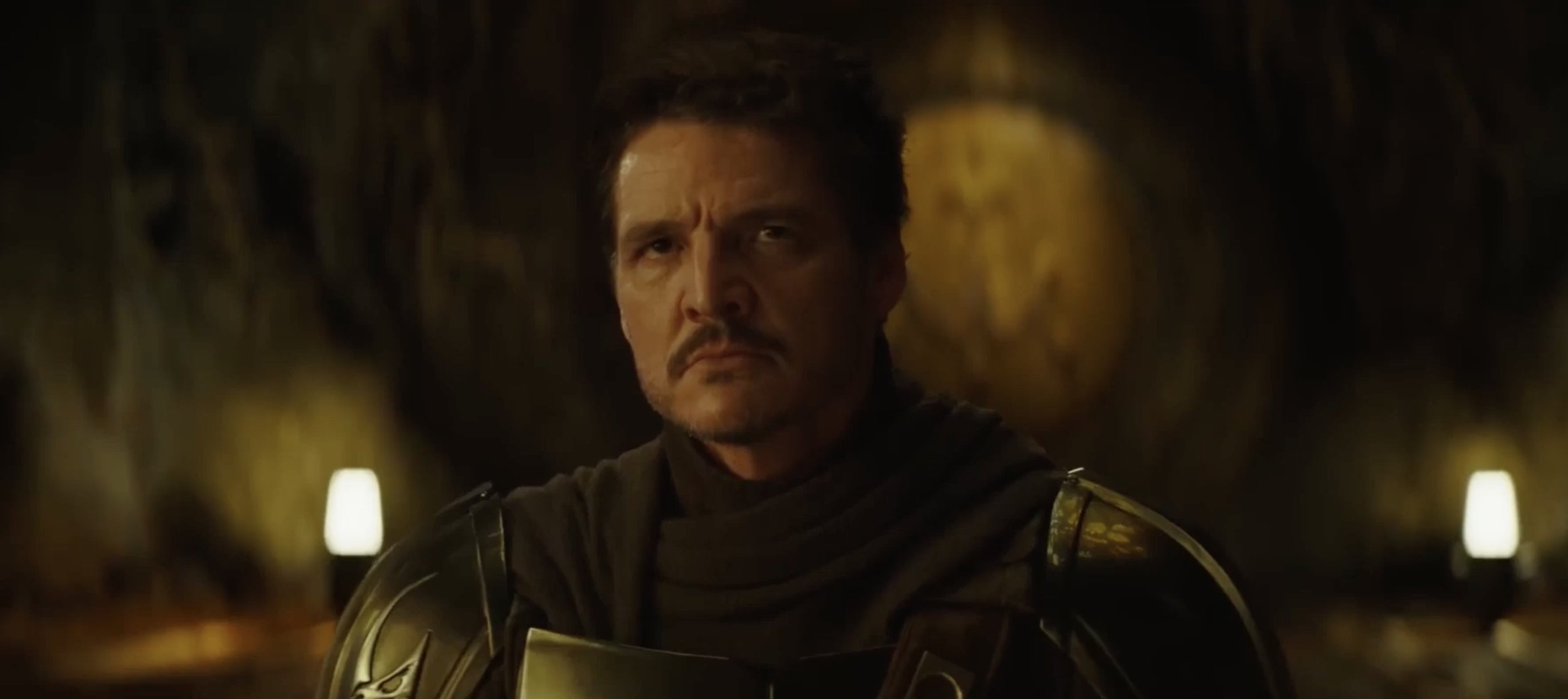 A man with dark hair and a mustache in a dark setting, wearing armor or a costume with metallic details, looking serious and intense.