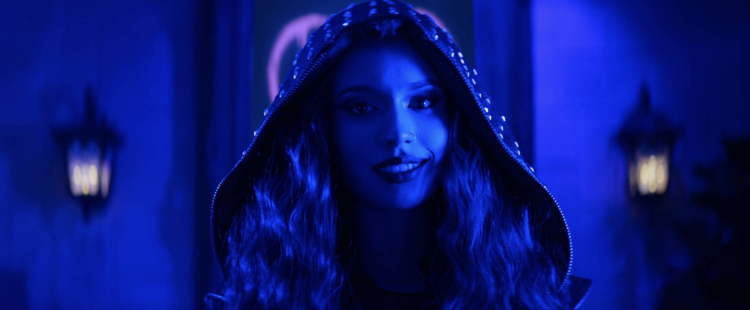 A woman with long curly hair wearing a hooded jacket, illuminated by blue lighting, standing in a corridor with lanterns on the walls.