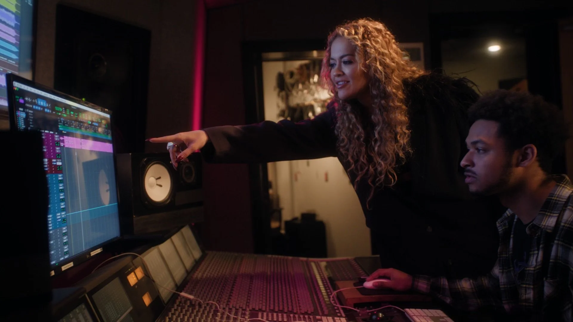 Two people working together in a music studio, one woman pointing at a digital audio workstation on a large computer screen, and a man sitting at a mixing console with headphones, surrounded by audio equipment.