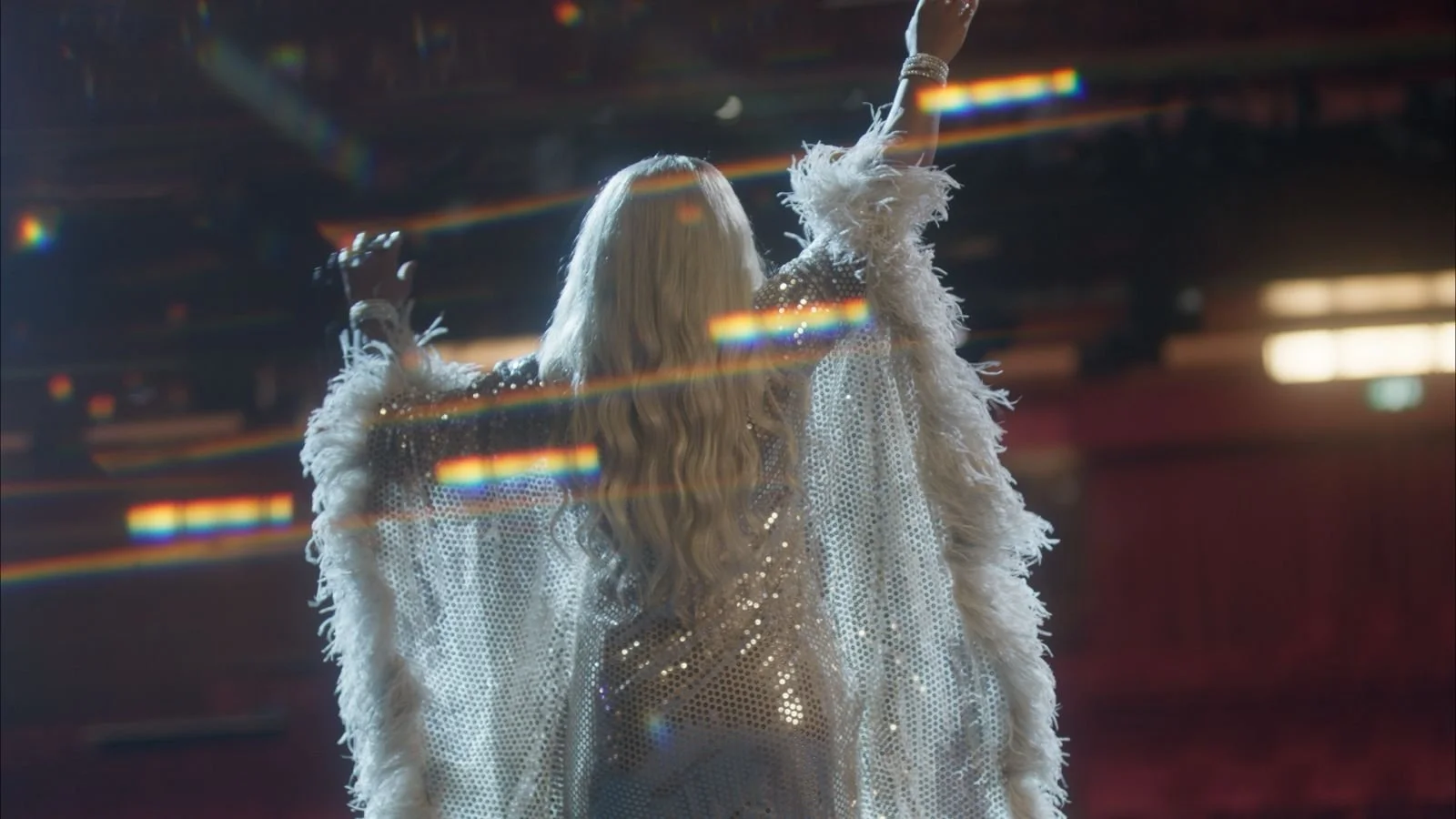 A performer with long wavy blonde hair dancing on stage, wearing a sparkling, feathered, and sequined white outfit, with colorful light reflections and a dark, blurred background.