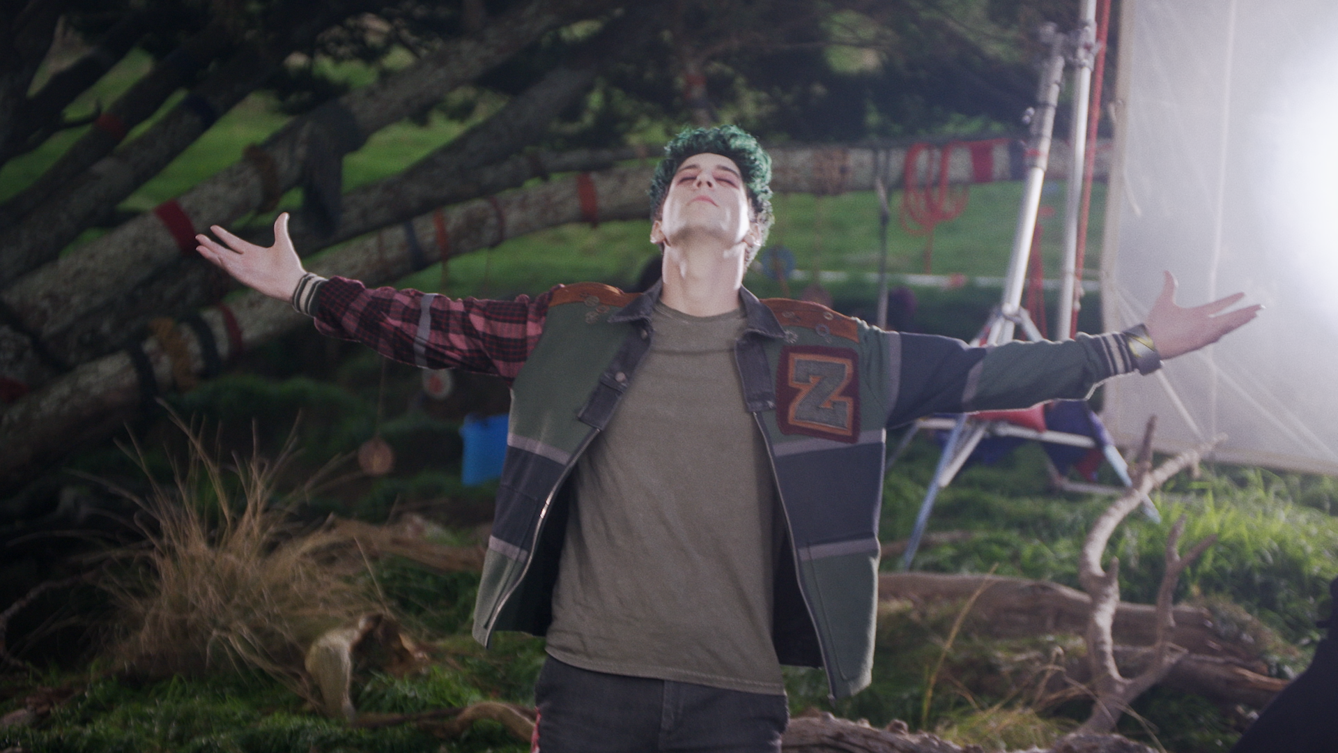 A young man with arms wide open standing outdoors, with a fallen tree and camera equipment in the background.