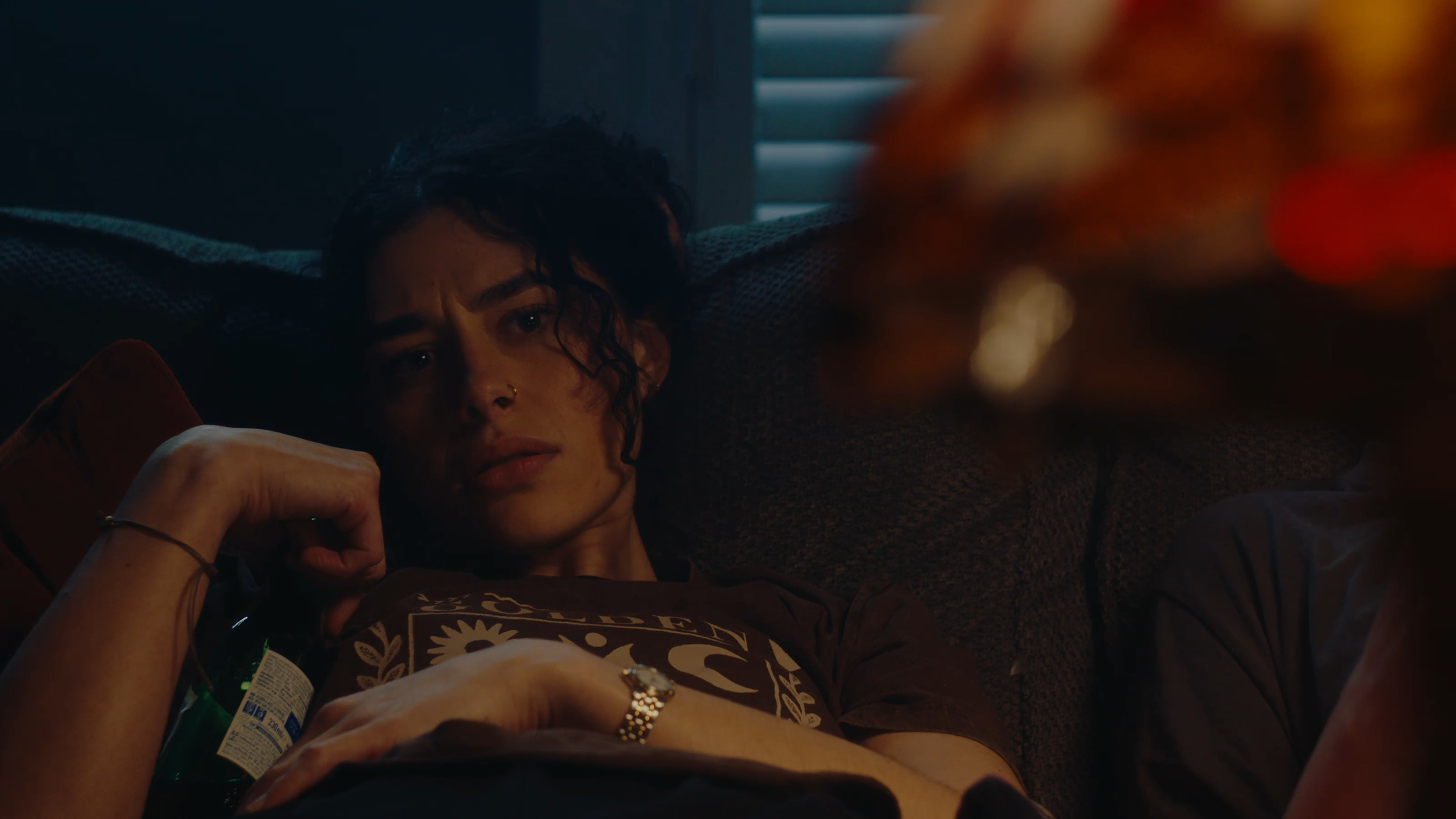 A woman with dark curly hair lying on a couch, resting her head on a cushion, looking thoughtful, with blurred lamp in the foreground.
