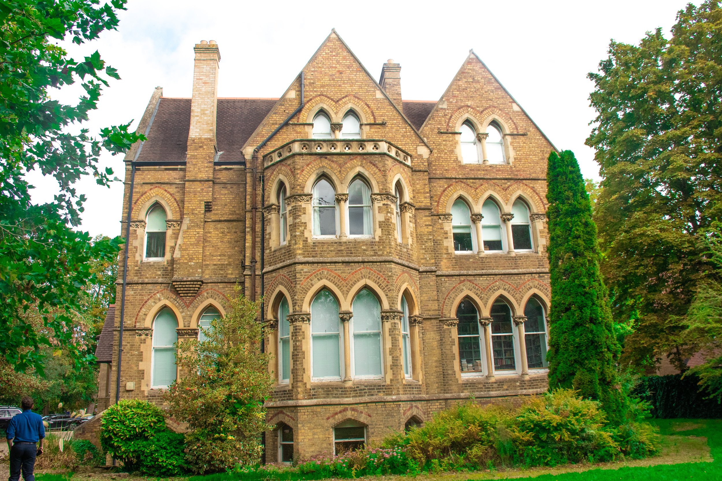 A historic brick building with Gothic-style arched windows, surrounded by greenery and trees.