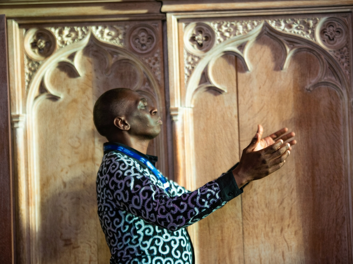 A man with dark skin and a patterned shirt is standing in front of an ornately carved wooden wall, with his hands raised in a prayerful or pleading gesture and eyes looking upward.