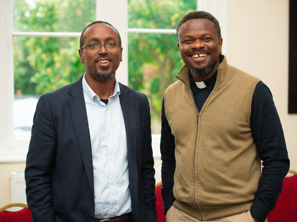 Two men smiling indoors near a window with green trees outside. One is dressed in a suit with a white shirt and glasses. The other is wearing a black shirt with a clergy collar and a tan vest.