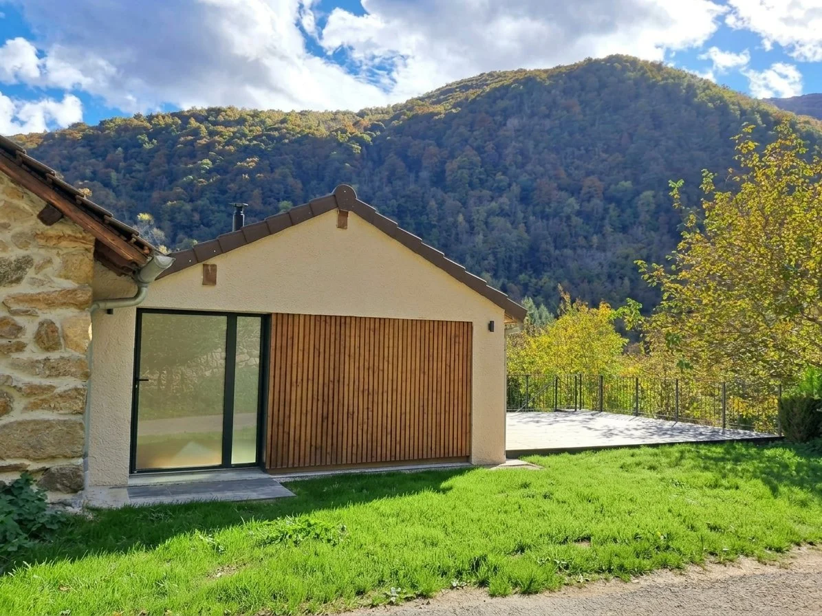 Maison avec vitre et porte en bois, entourée d'un jardin vert avec arbres, montagne en arrière-plan et ciel ensoleillé avec quelques nuages.
