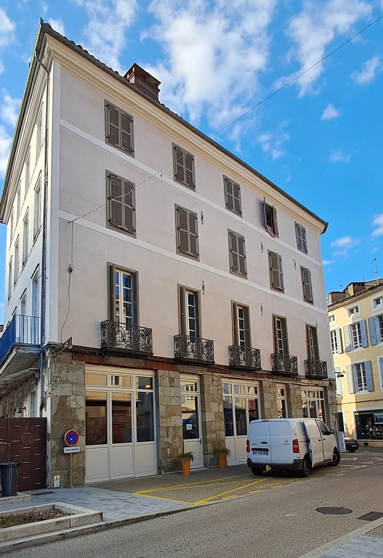 Immeuble à plusieurs étages avec fenêtres à volets en bois et balcons en fer forgé, ciel bleu avec quelques nuages.