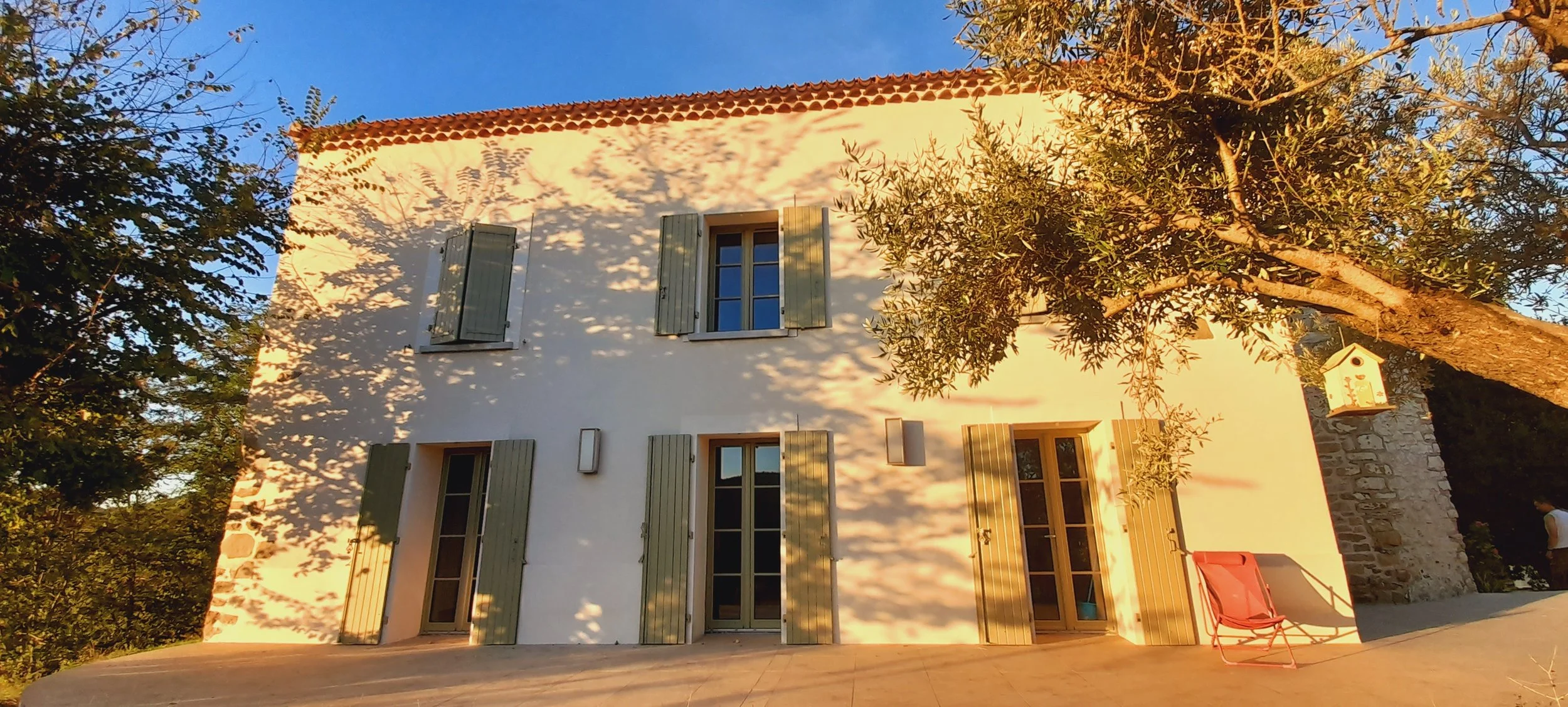 Maison blanche à deux étages avec volets verts, entourée d'arbres, lors du coucher du soleil.