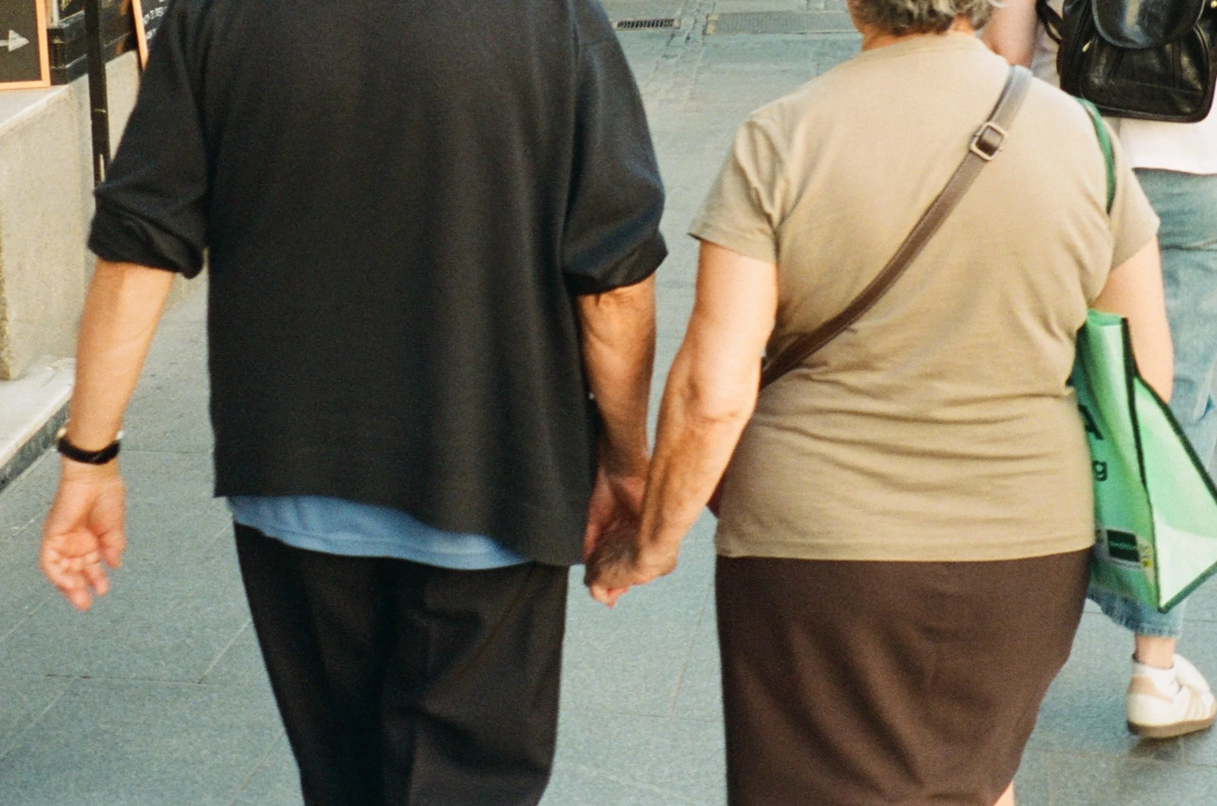 Two people walking hand in hand down a sidewalk, seen from the back, with other pedestrians in the background.
