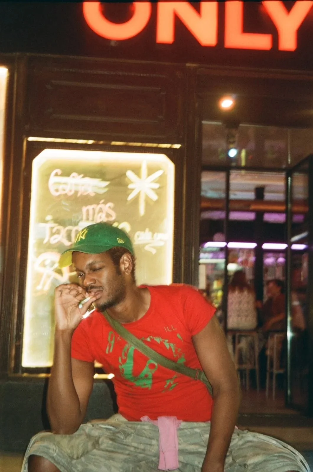 A young black man wearing a green cap and a red T-shirt sitting outside of a store at night, holding a cigarette near his face with a neon sign in the background.