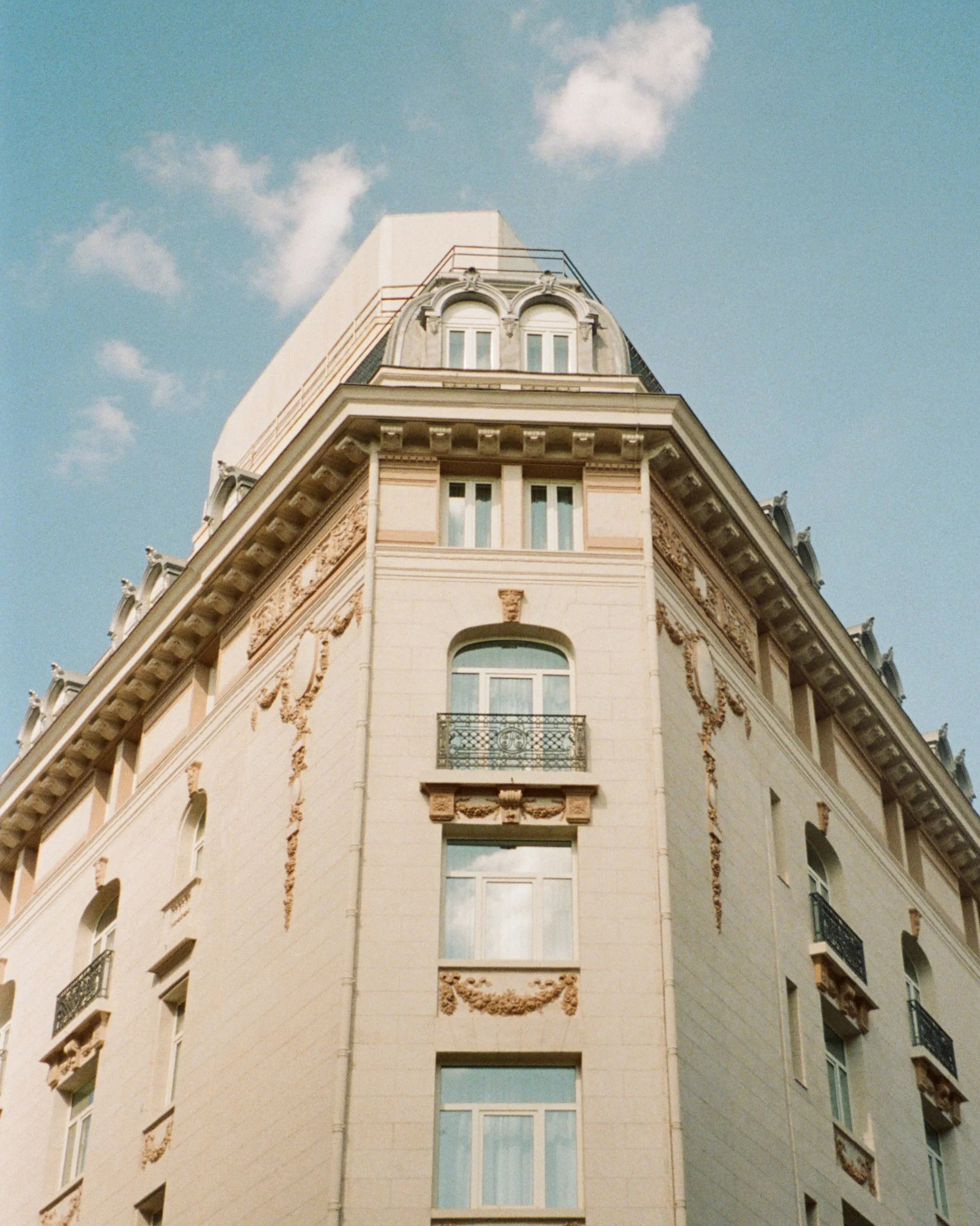 A tall building with ornate architectural details, including arched windows, decorative moldings, and a wrought iron balcony, against a blue sky with a few clouds.