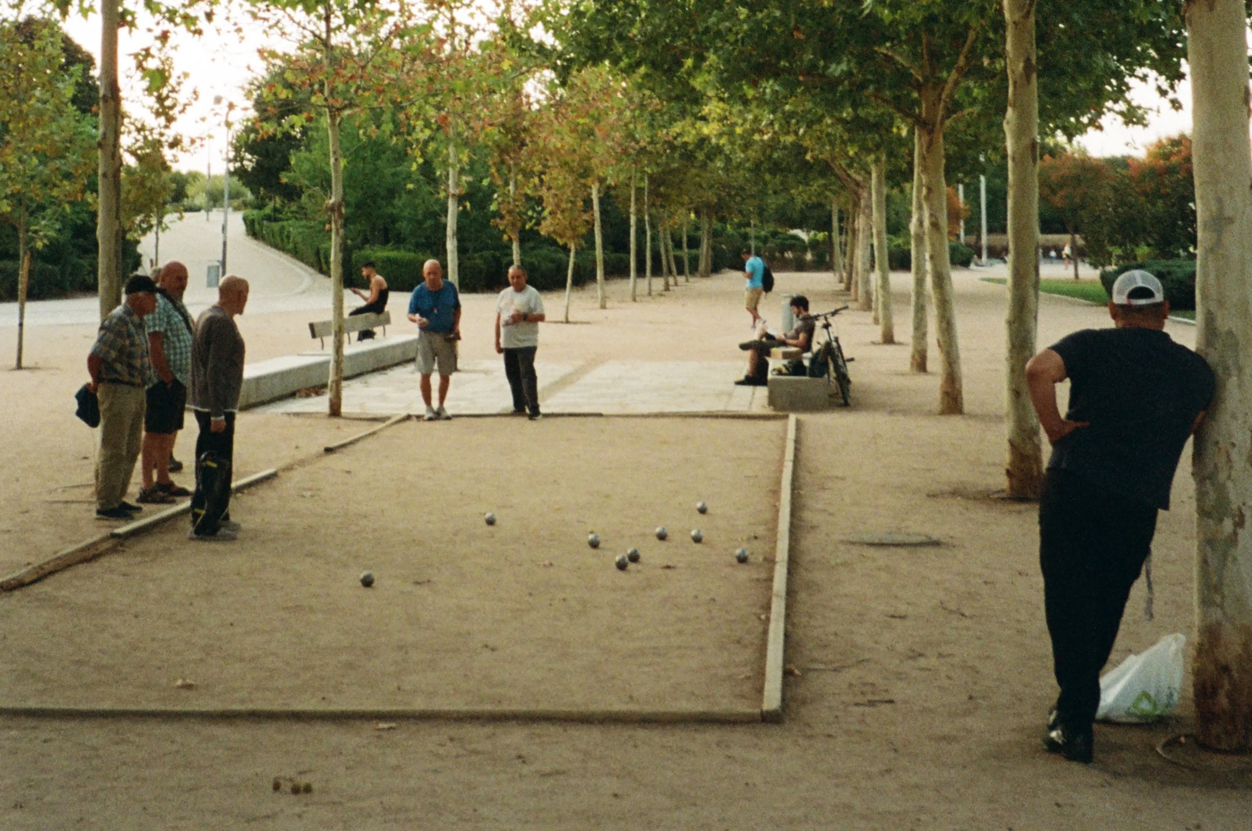 People playing pétanque in an outdoor park with trees and benches, some standing and some sitting, during daytime.