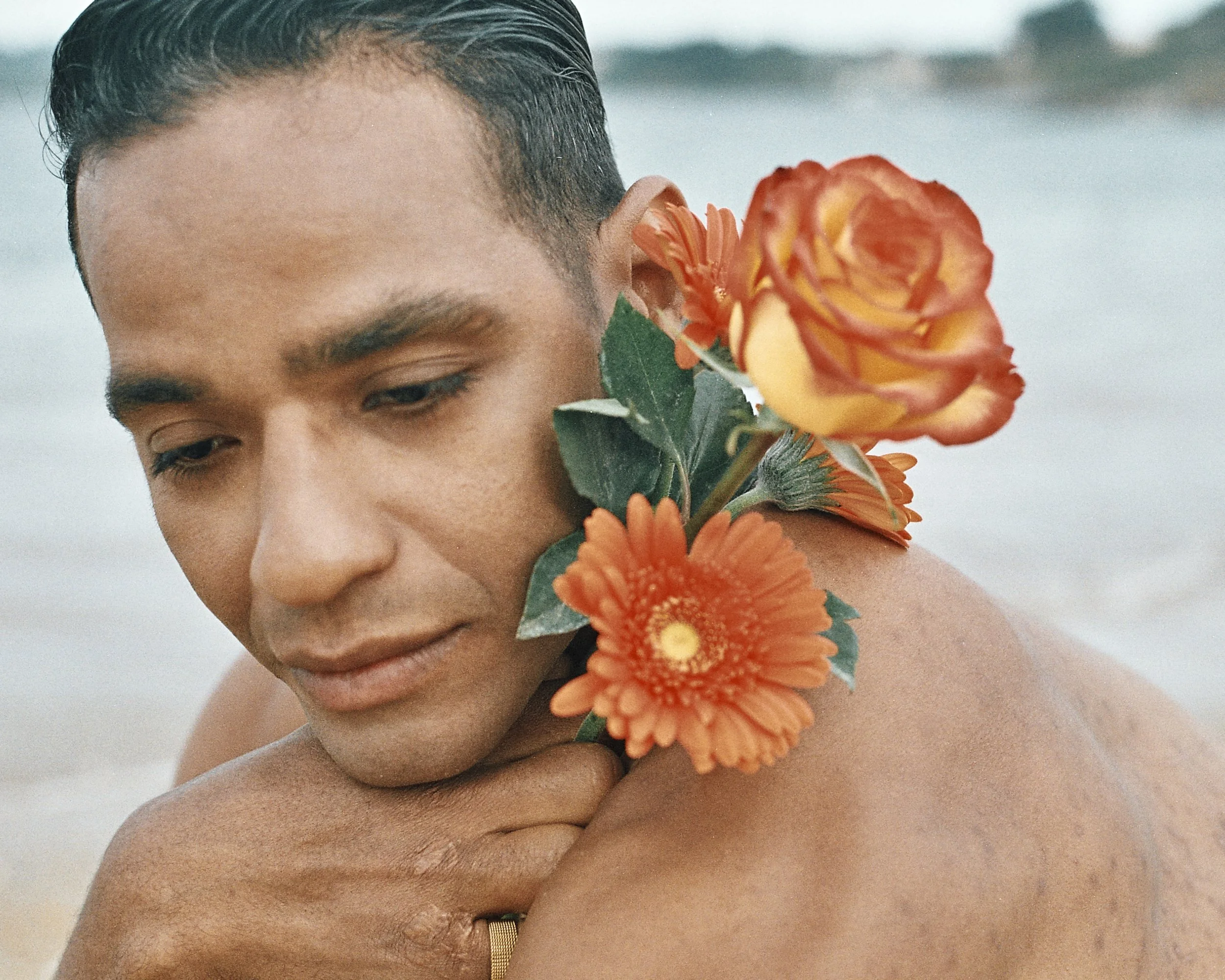 A person with short dark hair and medium skin tone holding orange flowers close to their face and shoulder, with a blurred sky or ocean background.
