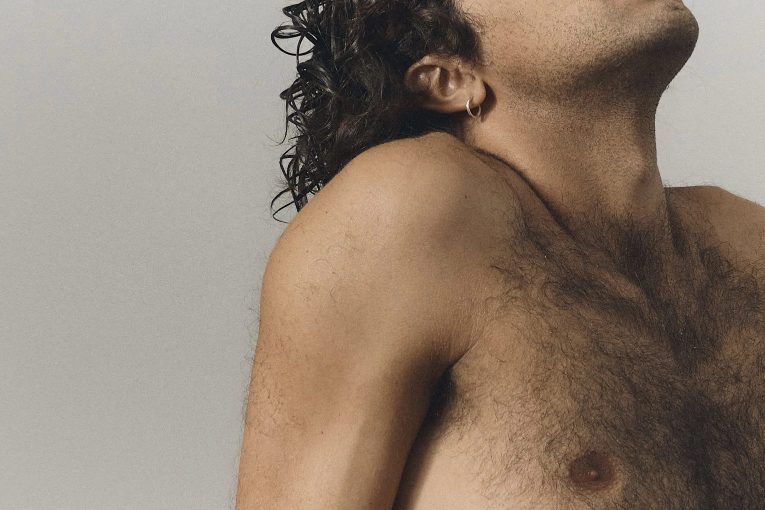 Close-up of a shirtless man with curly dark hair, wearing a hoop earring, and a light background.