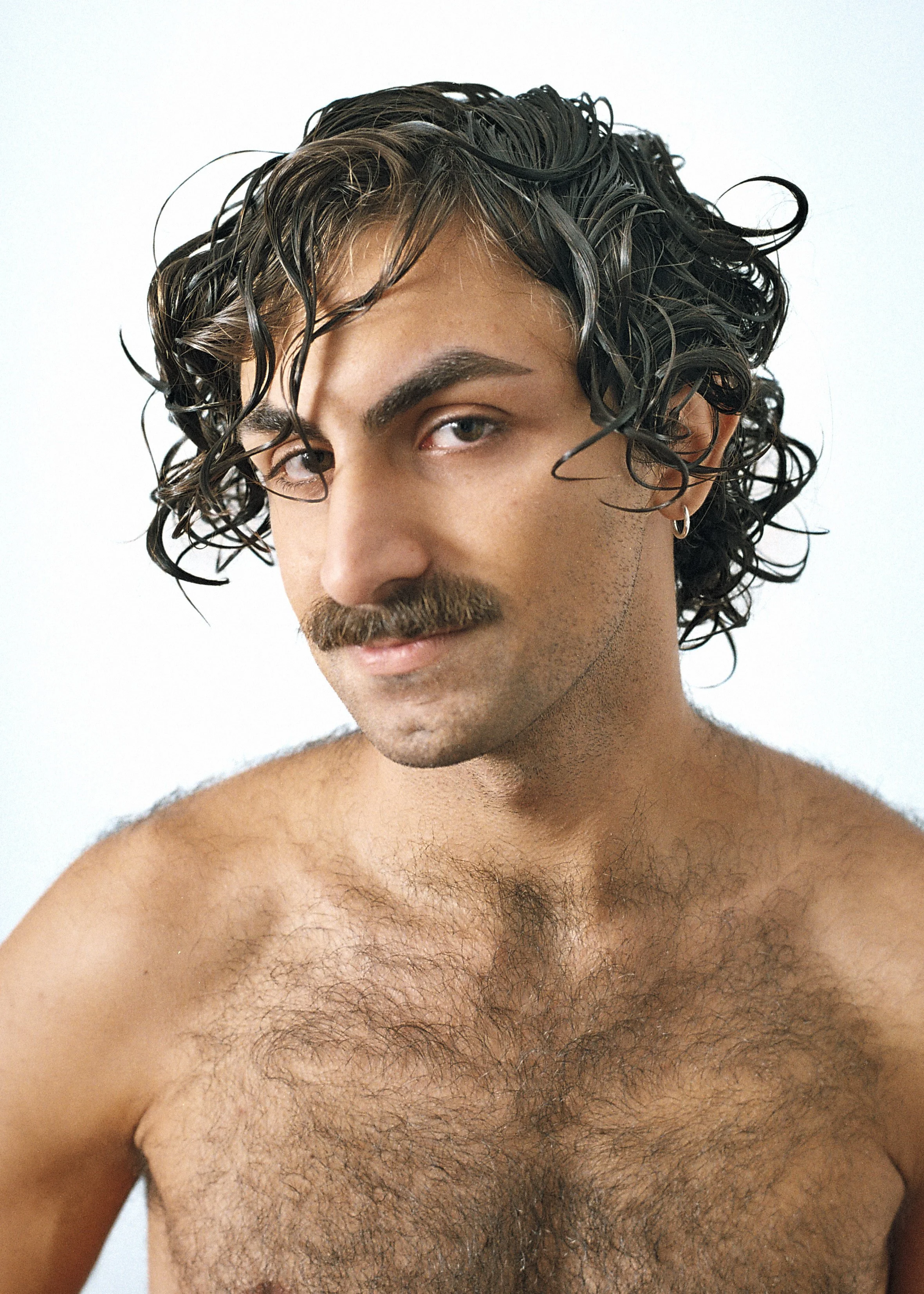 Close-up of a shirtless man with curly dark hair, a mustache, and chest hair, against a plain light background.
