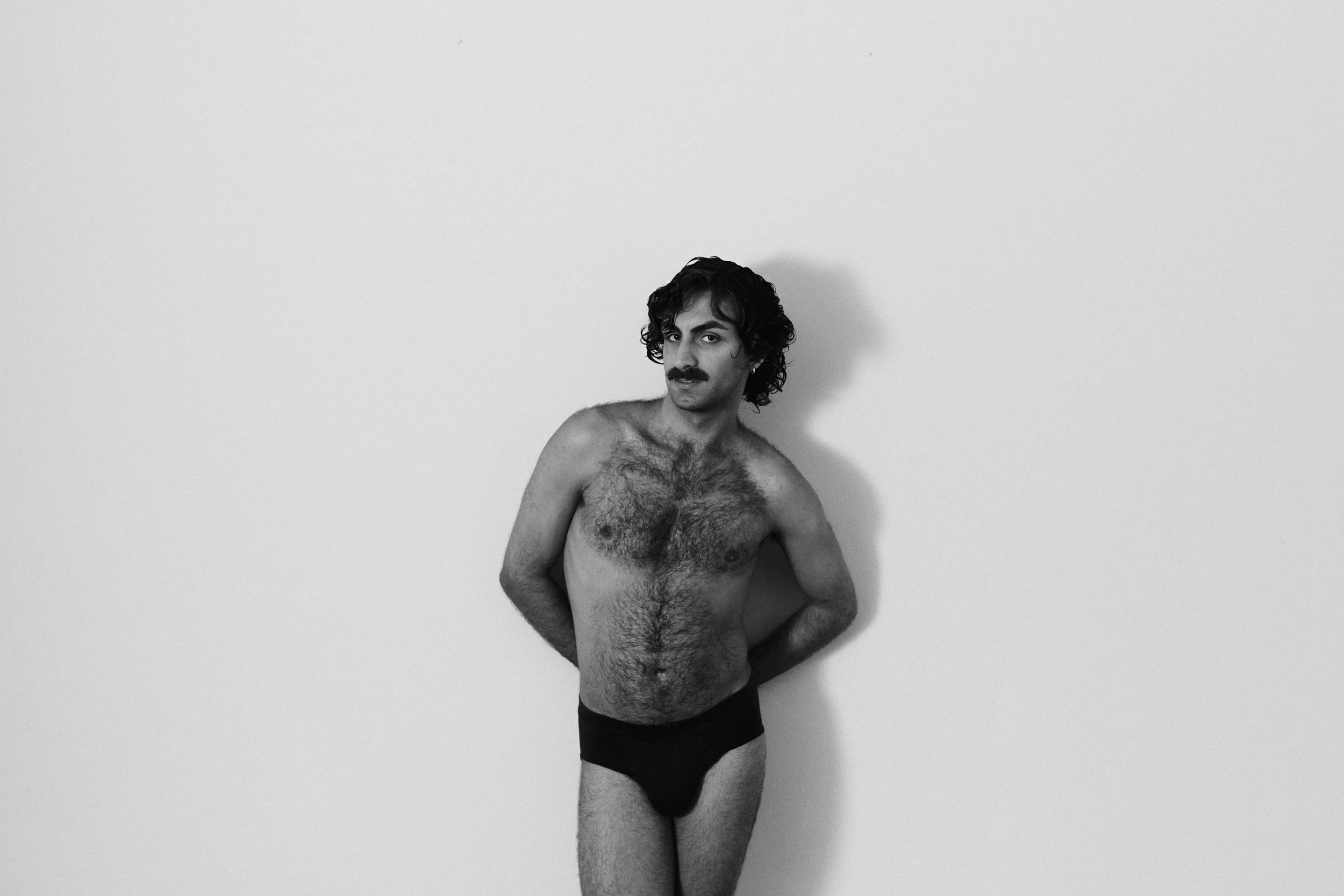 A black and white photo of a shirtless man with dark curly hair and a mustache, standing against a plain white wall. He is wearing black underwear and has his right hand resting on his waist.