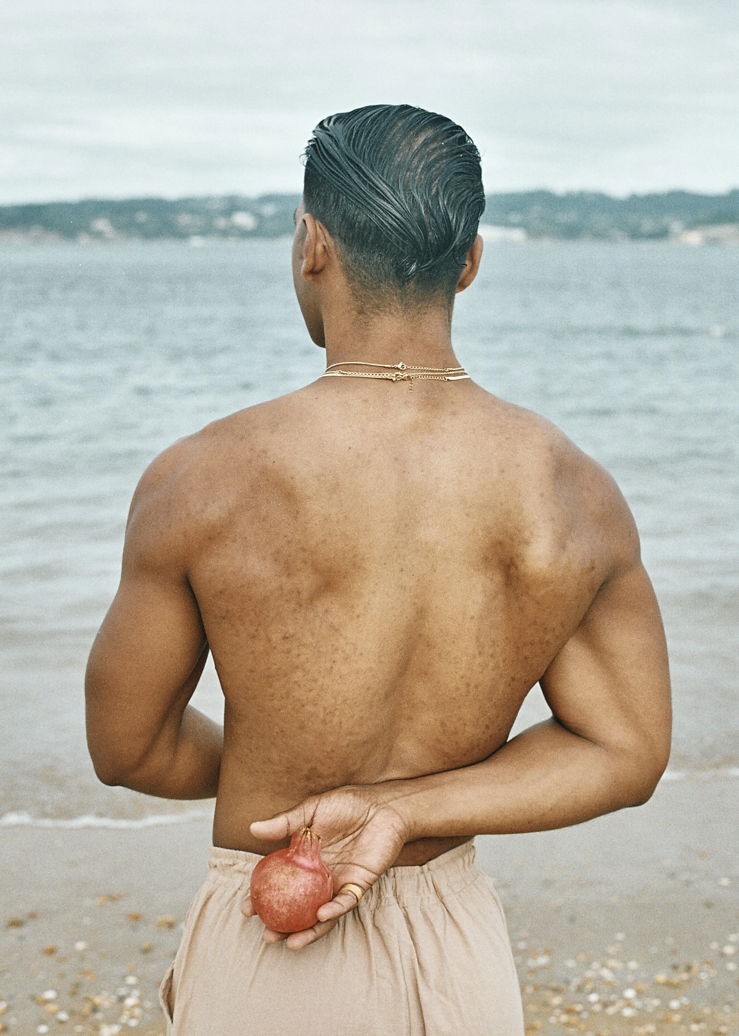 Person with wet hair, shirtless, holding a pomegranate behind their back, standing on a beach with water and cloudy sky in the background.