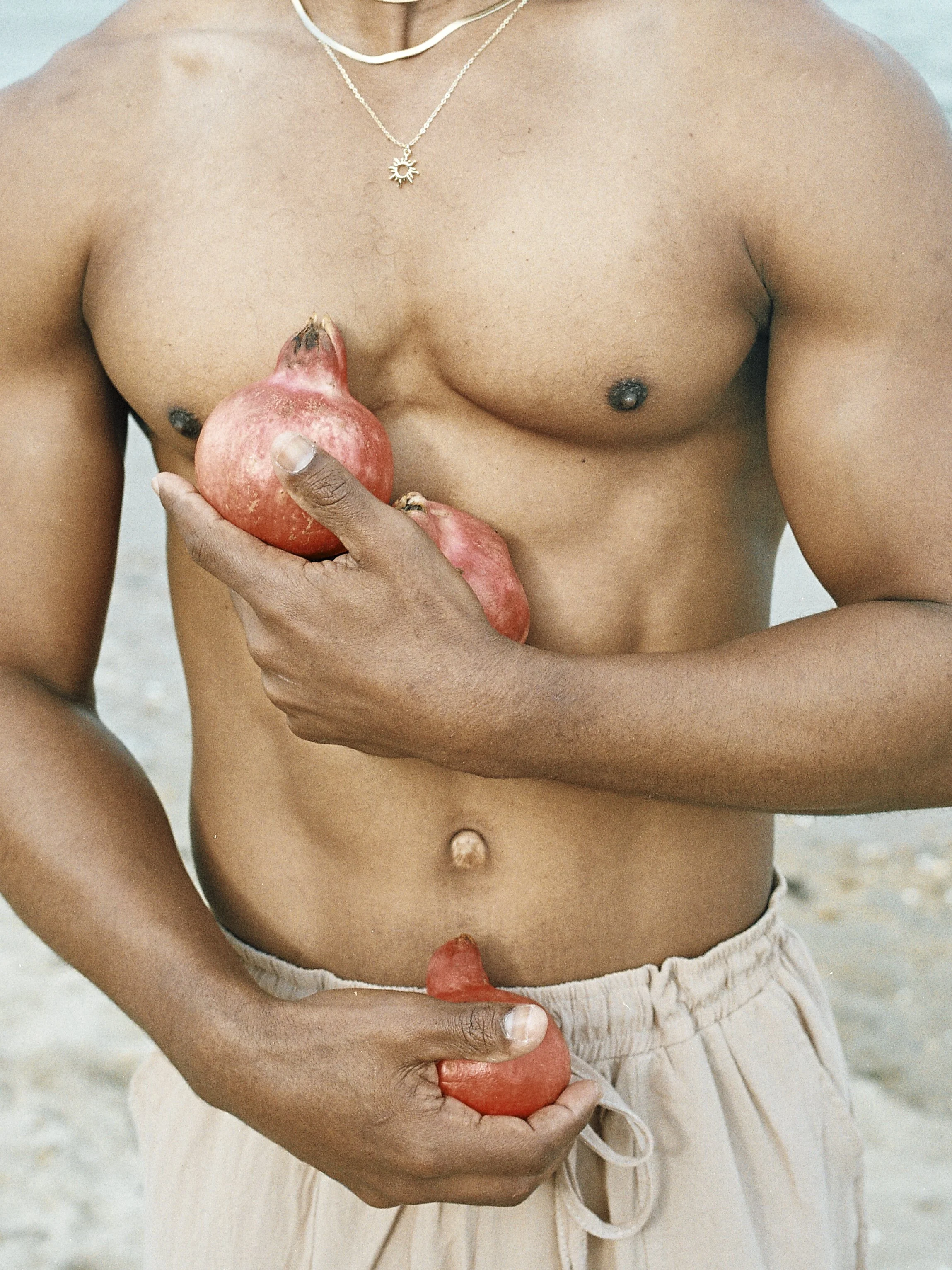 A person with medium brown skin is standing shirtless on a sandy beach, holding a watermelon and a piece of watermelon, with the ocean or water in the background. They are wearing a necklace with a sun pendant.