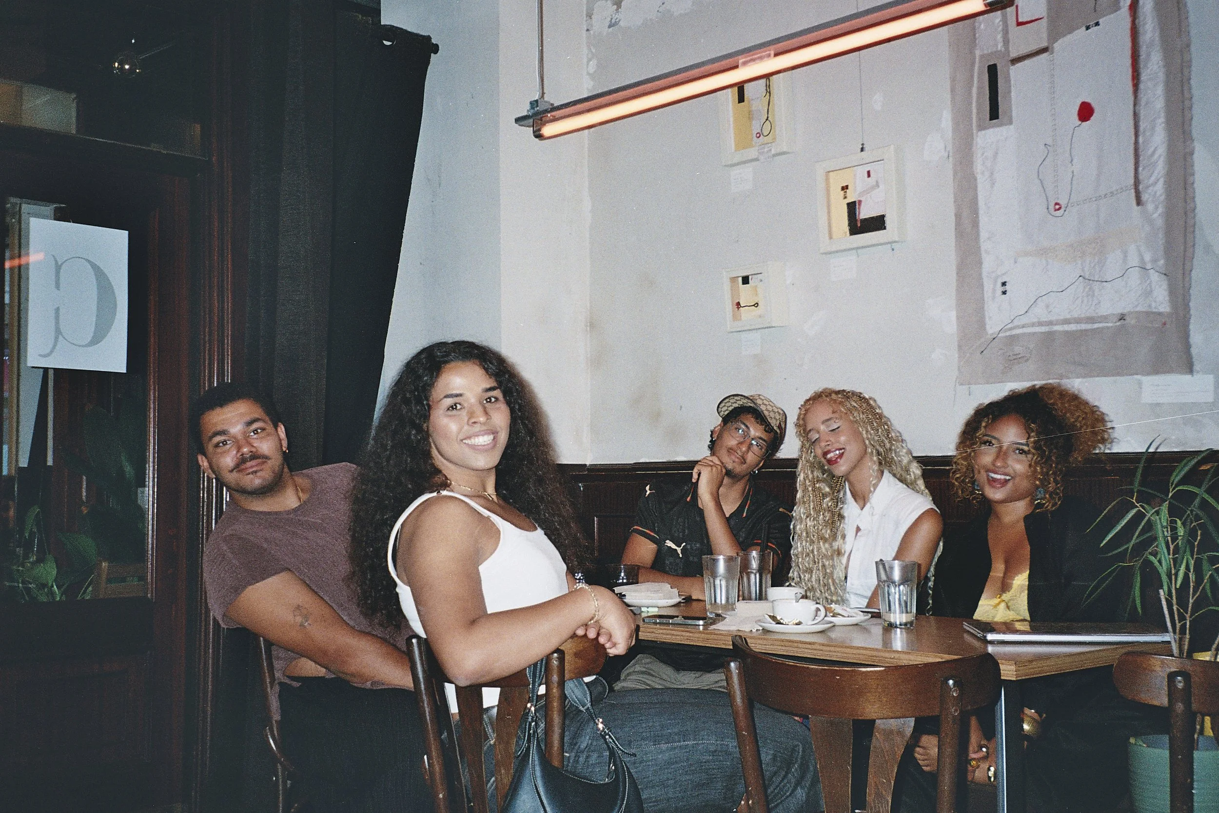 Group of five young adults sitting around a dining table in a restaurant, smiling and enjoying their time together. The table has plates, glasses, and a smartphone. The background features a wall with framed artwork and a red neon light.