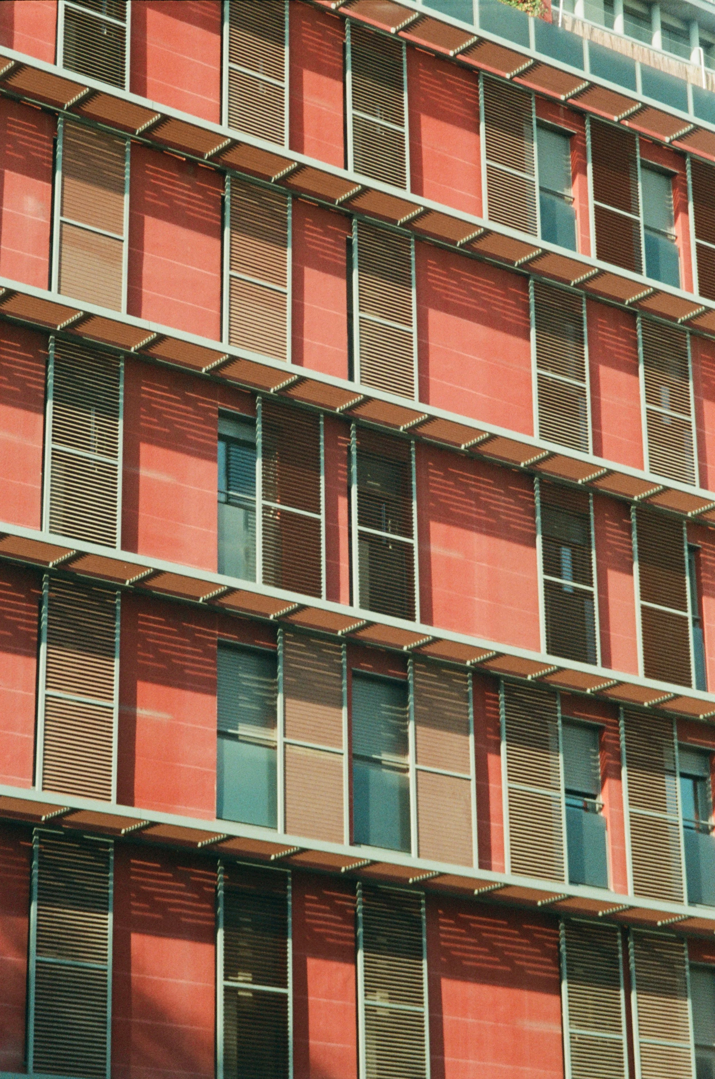 Close-up of a modern building facade featuring multiple floors with rectangular windows and brown louvered shutters, red exterior walls, and shadows cast by the shutters.
