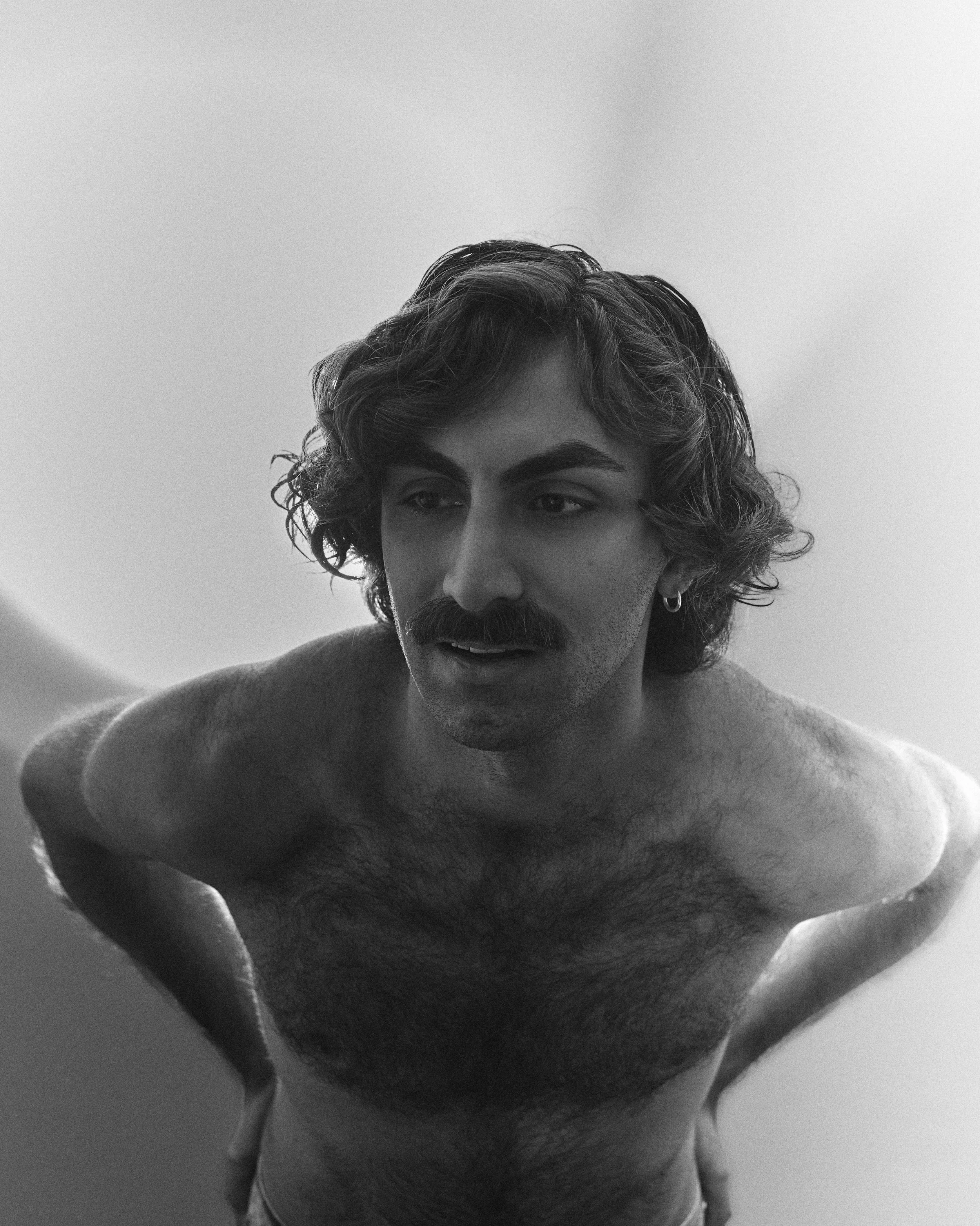 A black and white photo of a shirtless man with curly hair and a mustache, looking downward, with a plain background.