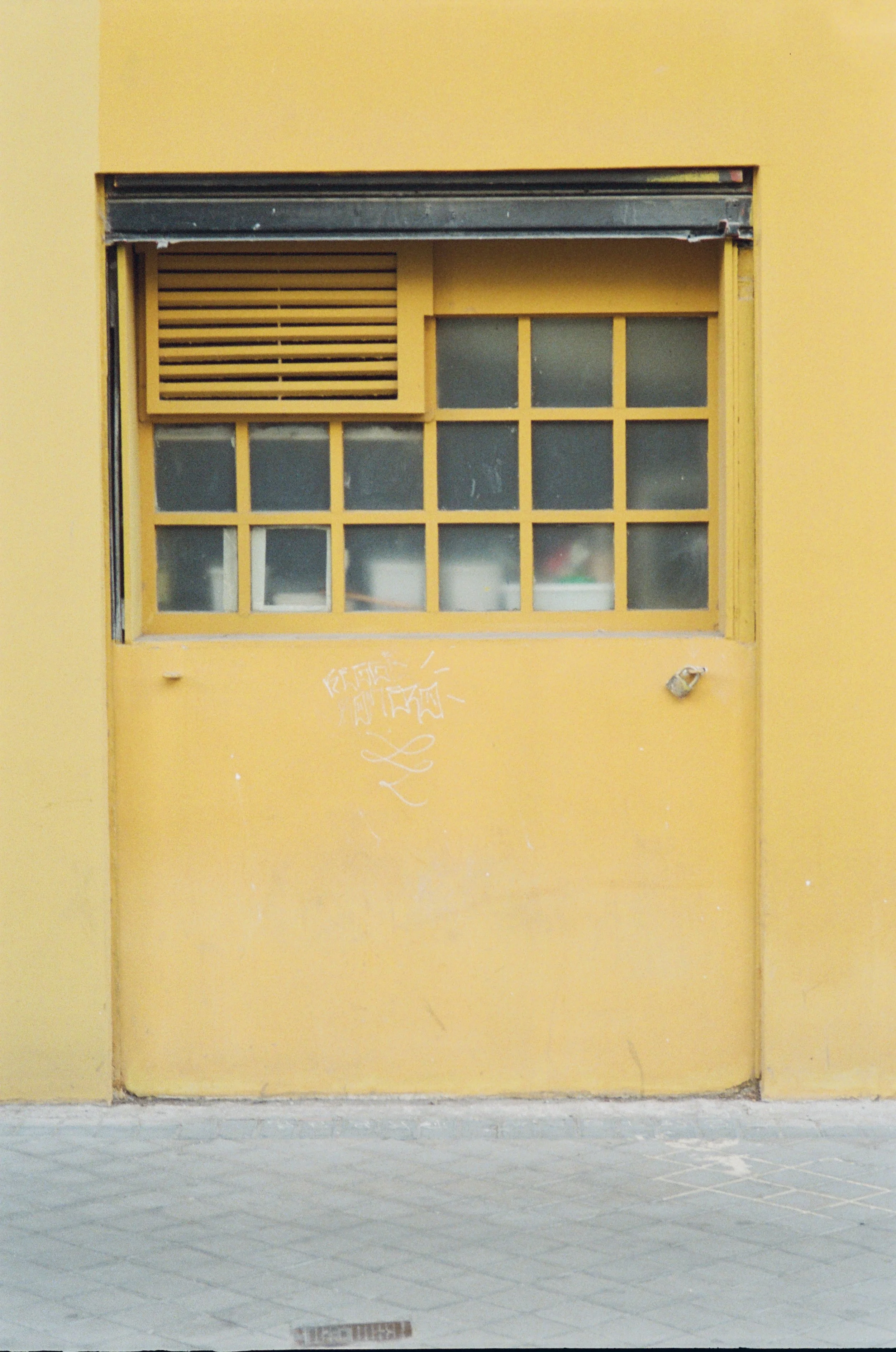 Yellow building wall with a yellow window divided into smaller panes and a yellow vent above it, with some white graffiti and markings on the wall.