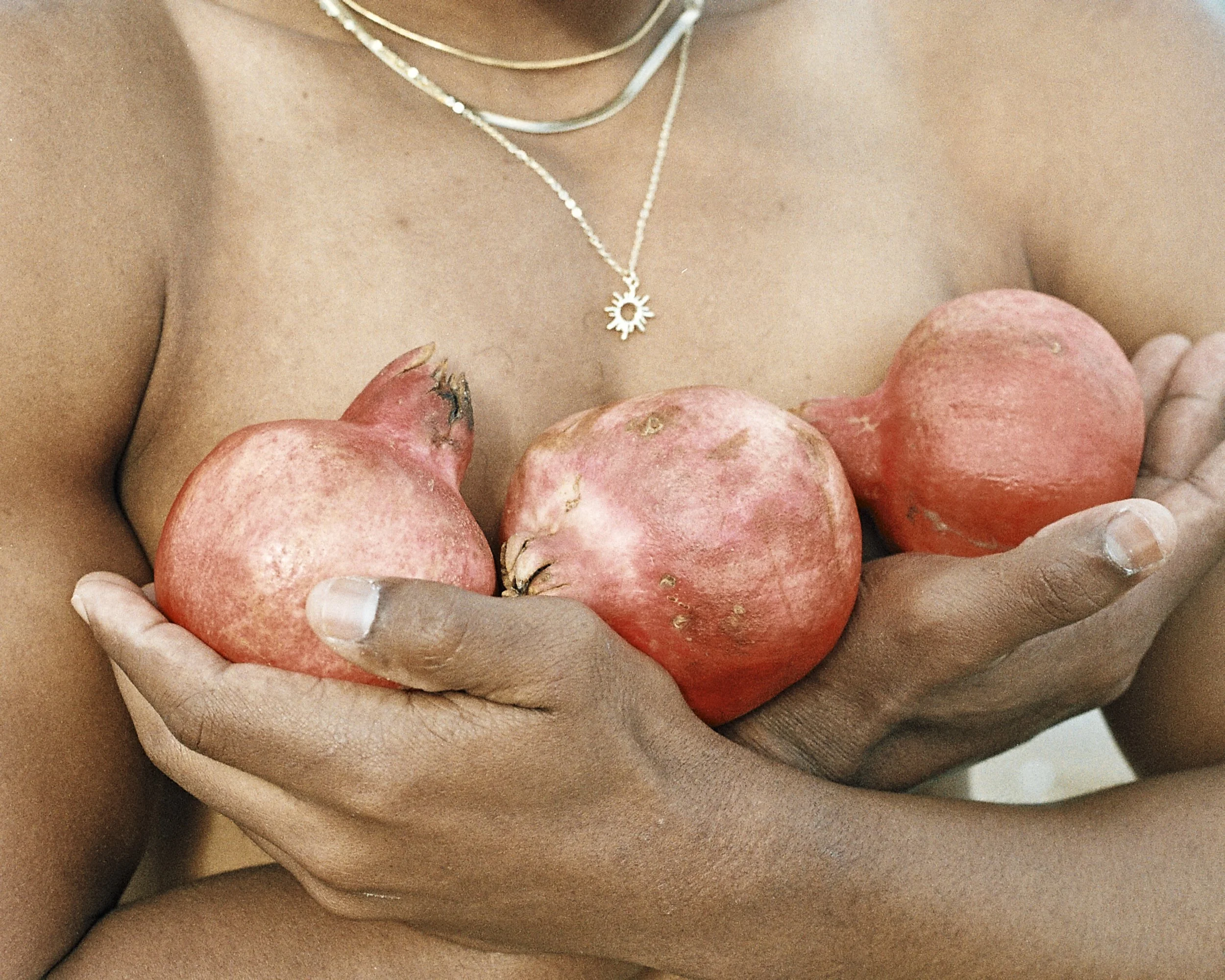 Close-up of a woman's torso with multiple red potatoes held in her hands, wearing a necklace with a sun-shaped pendant.