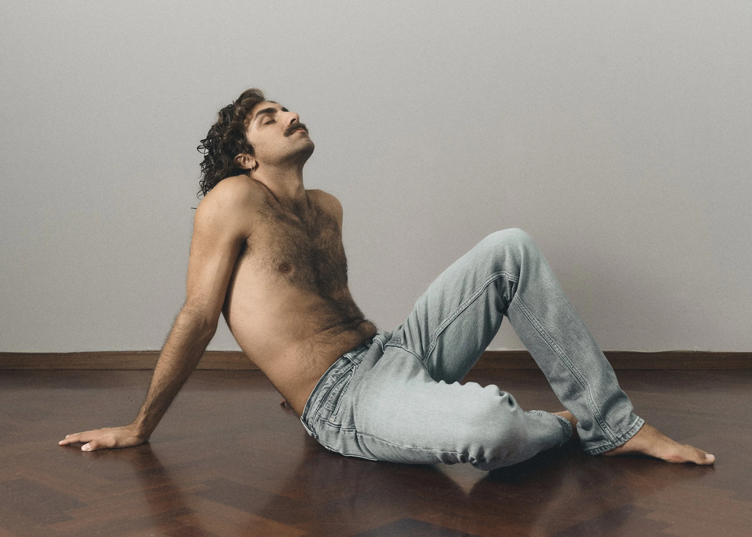 A man with curly hair and a mustache sitting on a wooden floor with his back against a wall. He is shirtless, wearing light blue jeans, with his head slightly tilted back and eyes closed.