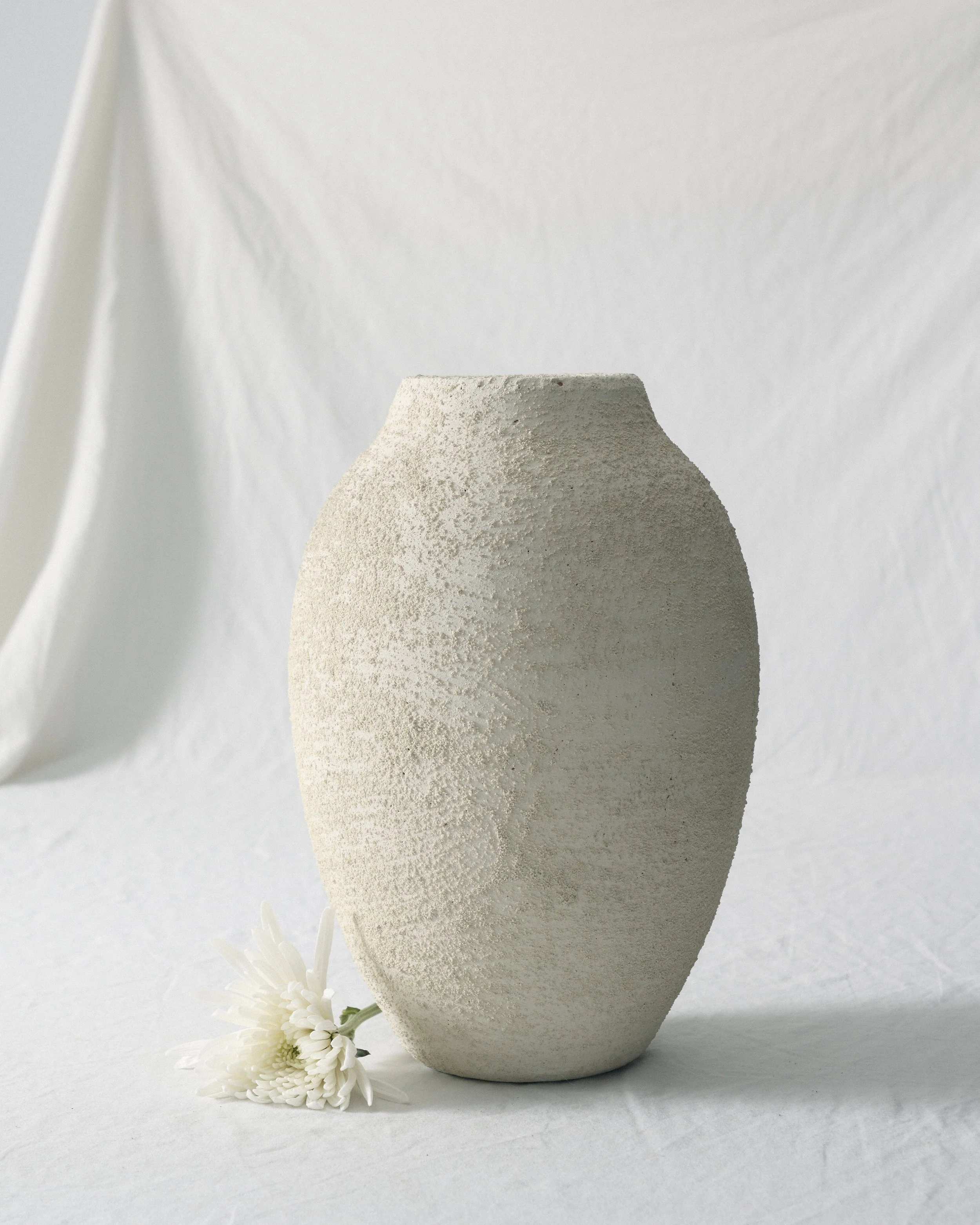 A white textured ceramic vase with a narrow opening, decorated with white flowers and green foliage, set against a white background.
