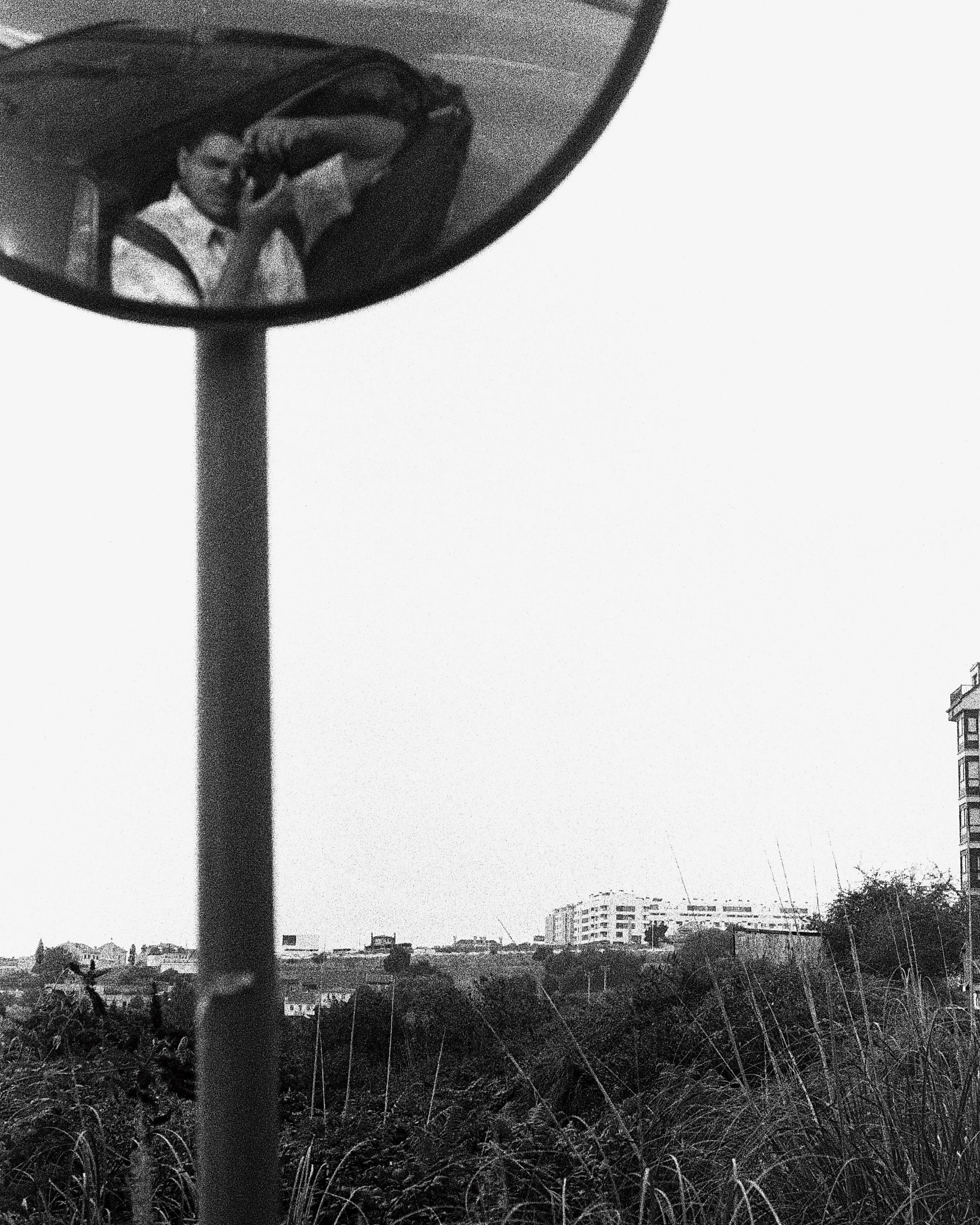 Black and white photo of a street lamp, with city buildings and a partly cloudy sky in the background. The foreground has tall grass or weeds.