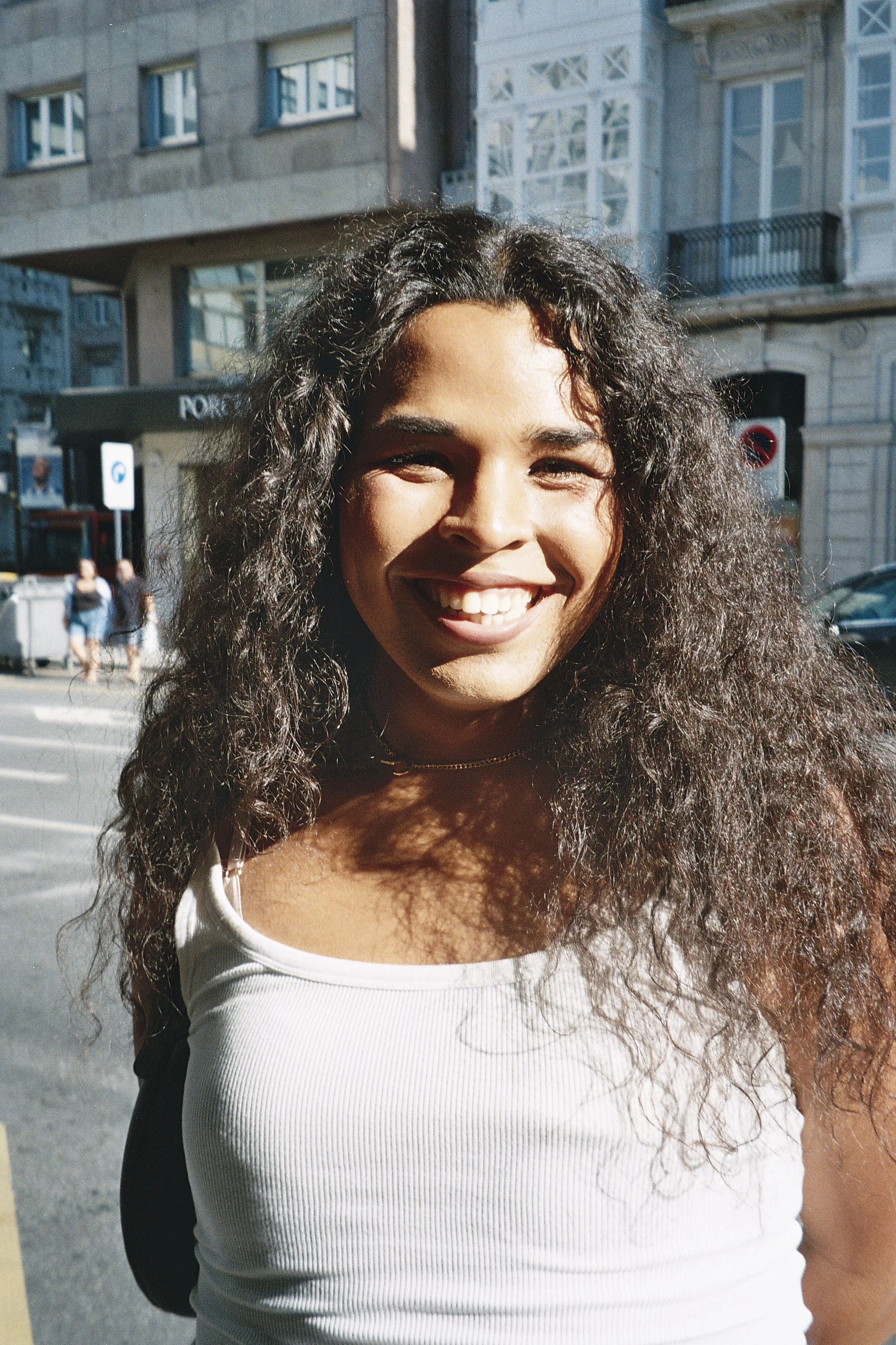 A black woman with curly hair smiling outdoors in a city street on a sunny day, wearing a white tank top.