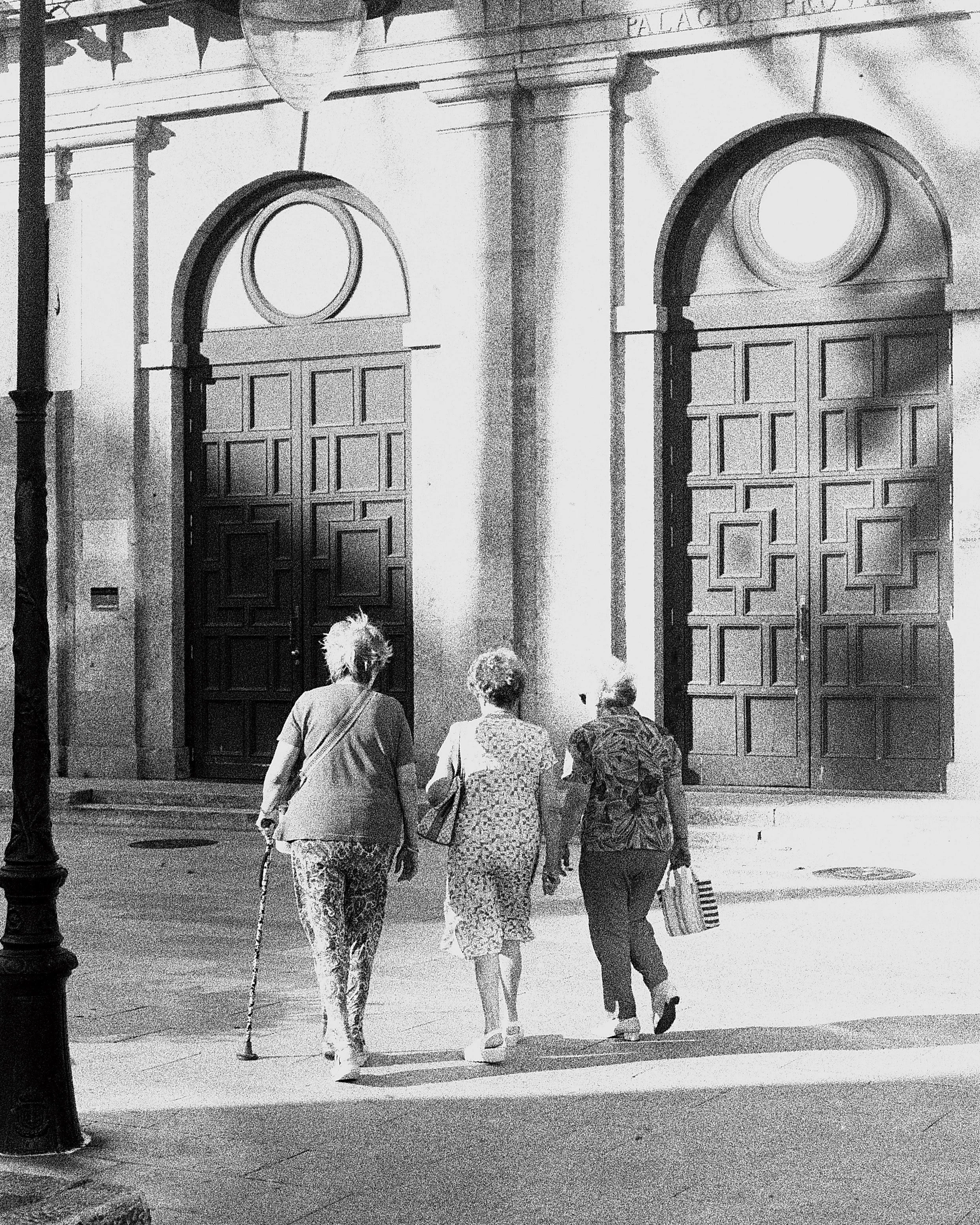 Four women walking in front of a large wooden door with two circular windows, carrying bags, and a lamppost visible on the left, with the word 'PALACIO' inscribed above the door.