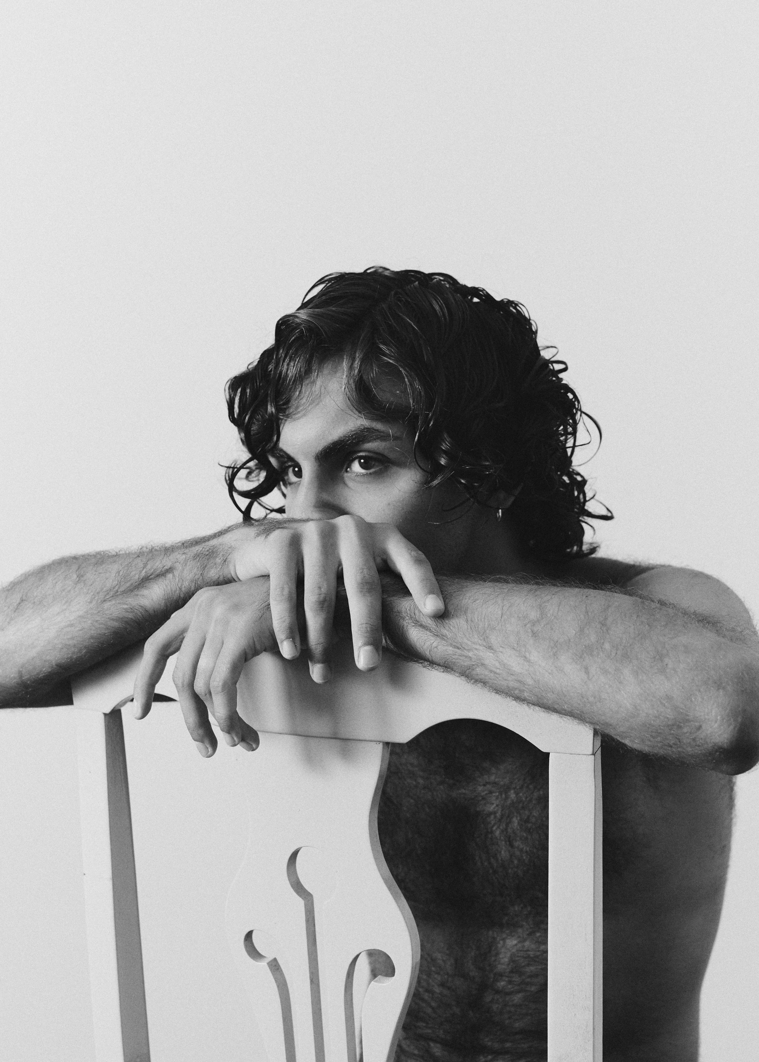 Black and white portrait of a person with curly hair sitting behind a wooden chair. The person's head and eyes are visible, with hands resting on the chair's top. The person's gaze is directed towards the camera.