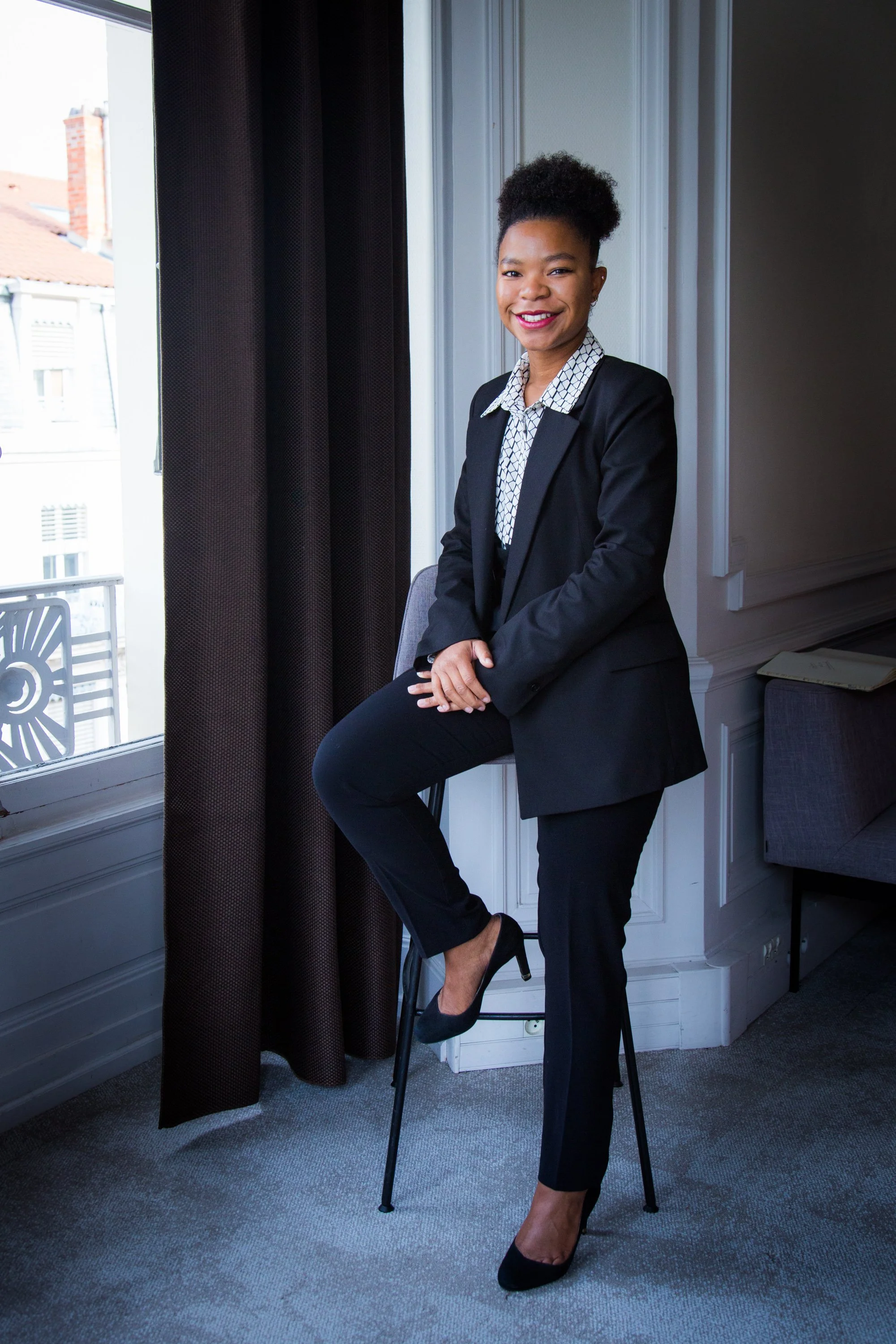 Une femme en tailleur noir avec une chemise à motifs noirs et blancs, assise sur une chaise près d'une fenêtre, souriant, dans une pièce lumineuse aux murs blancs.