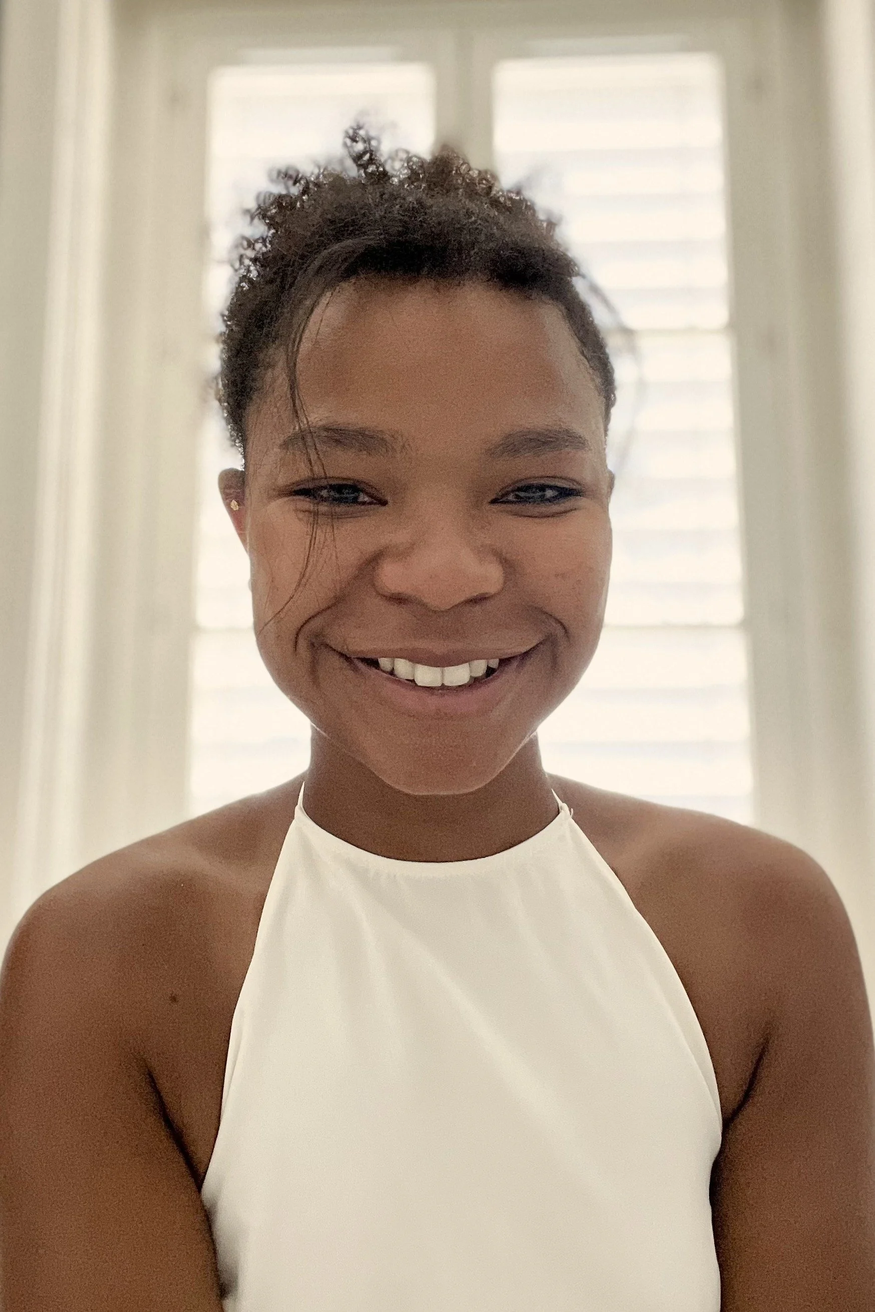 Une femme souriante portant une robe blanche, avec des cheveux courts bouclés, devant un fond clair avec deux fenêtres ou volets en arrière-plan.