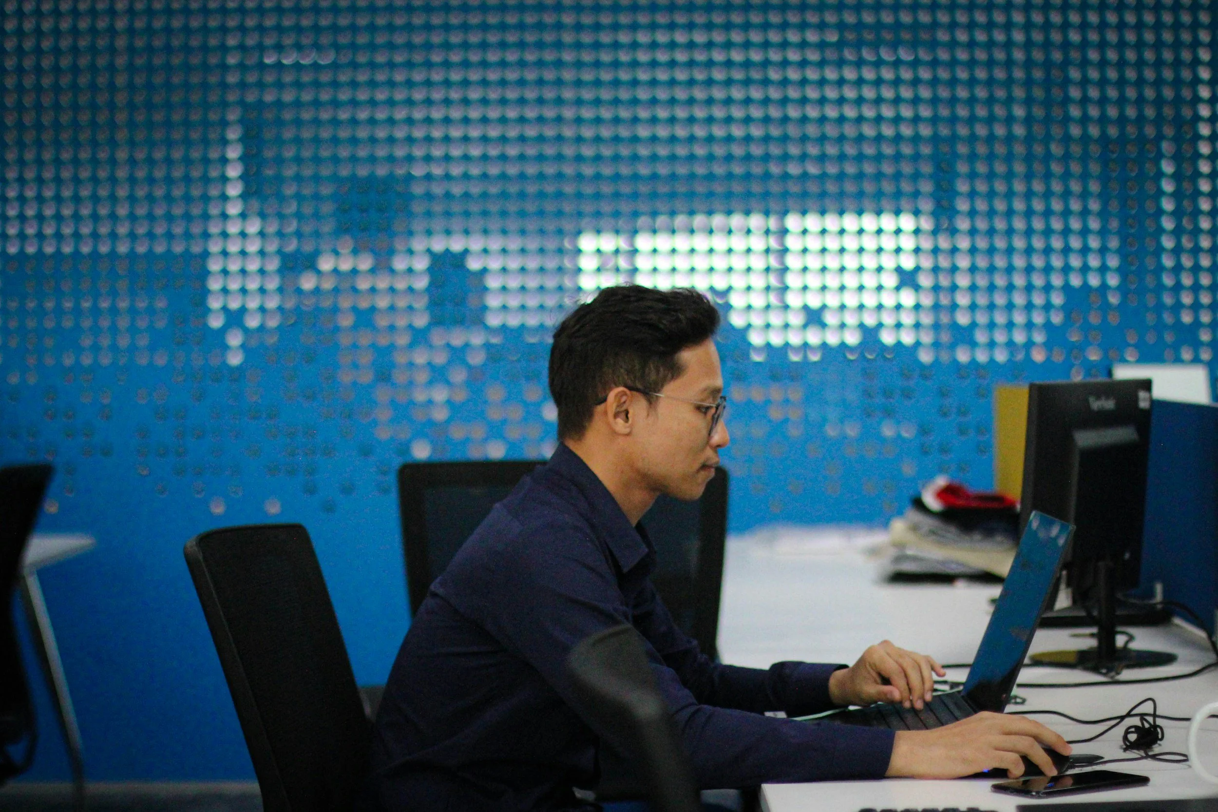 A man with black hair and glasses working on a laptop at a desk in an office with a blue dotted wall in the background.