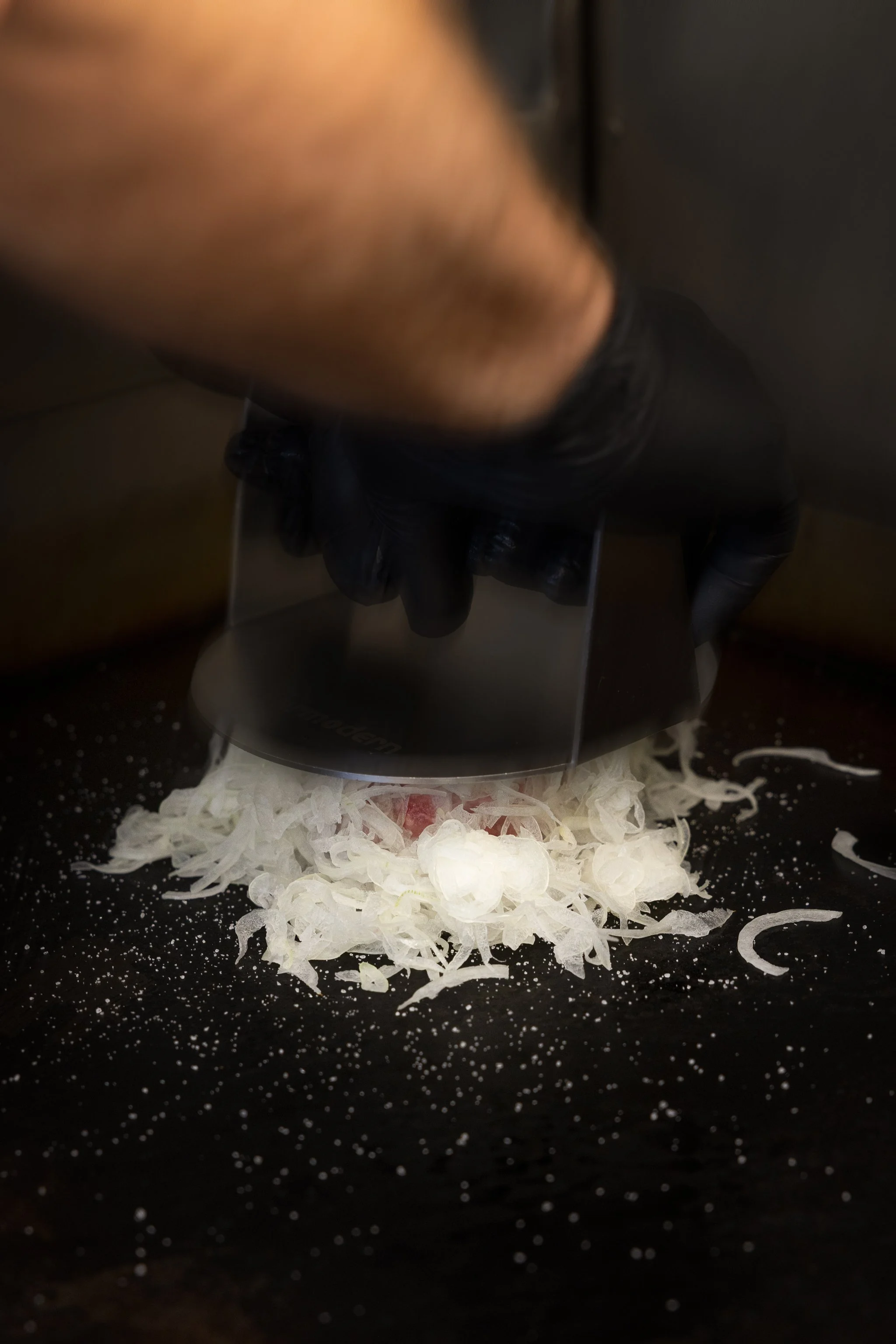 Nahaufnahme eines schwarzen Schneidebretts mit gehackter Zwiebel und einer Küchenmaschine, die die Zwiebeln schneidet, mit verstreutem Zwiebelmaterial auf dem Brett.