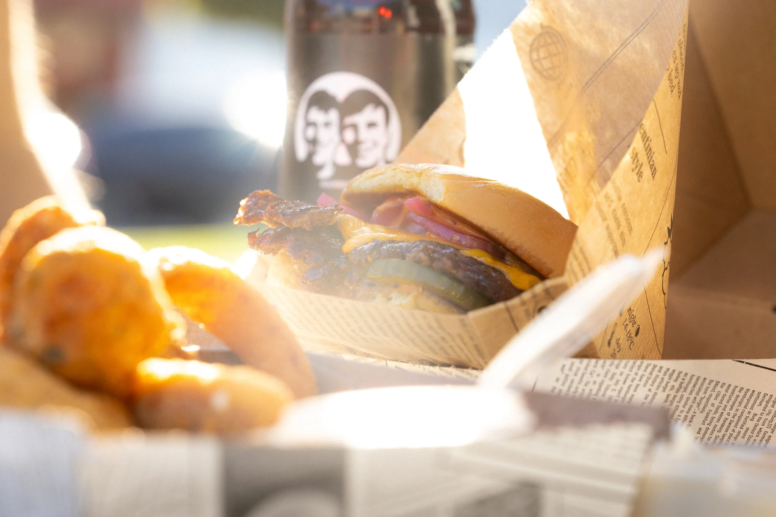 Ein Burger in einer Papiertüte, mit Fleischpatty, Käse, Salat, Tomaten und Zwiebeln. Im Hintergrund eine Flasche mit einem schwarzen Etikett.