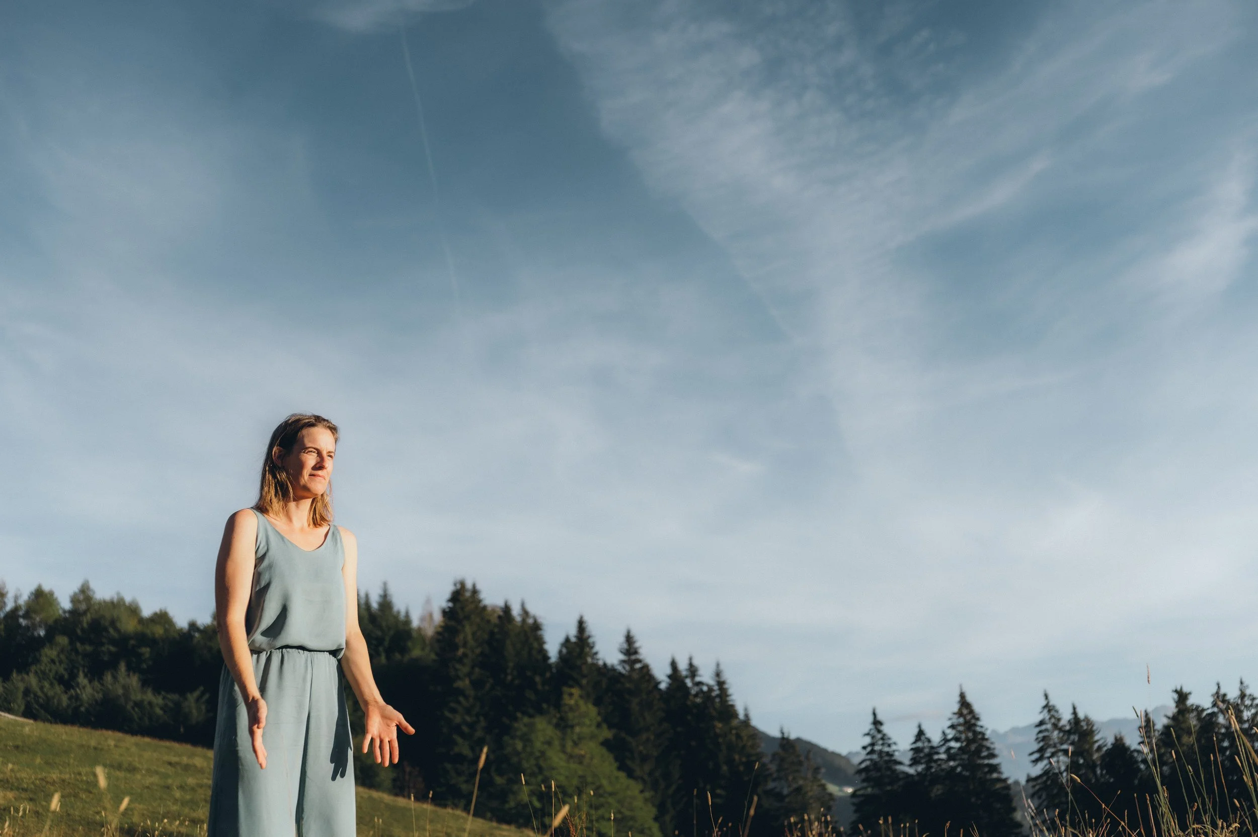 Eine junge Frau steht auf einer grünen Wiese mit Bäumen im Hintergrund und einem blauen Himmel mit Wolken und Flugzeugmerkmale. Sie trägt ein graues Kleid und scheint nach oben zu schauen.