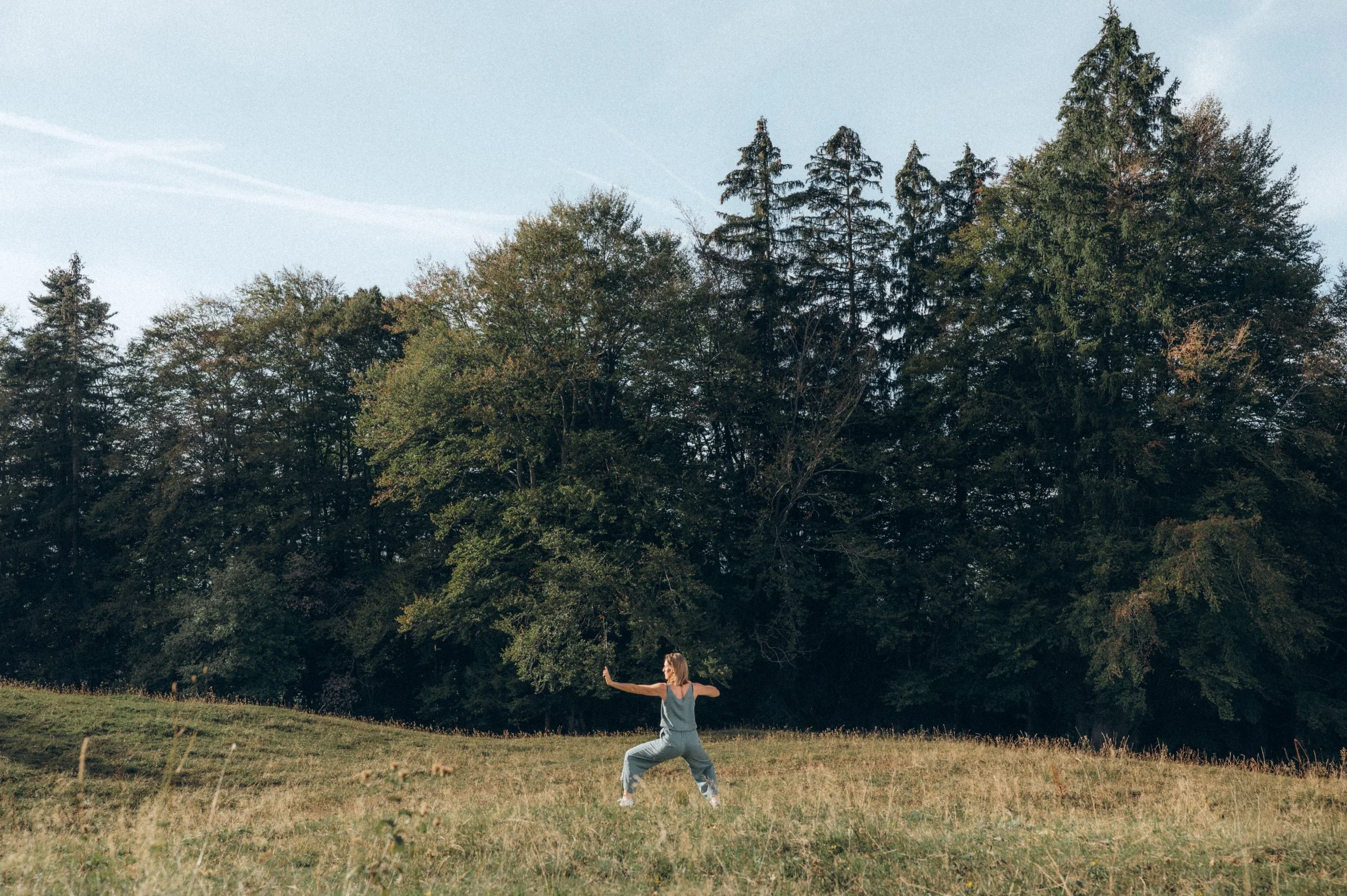 Eine Frau macht Yoga im Freien auf einer Wiese vor einem Wald mit hohen Tannenbäumen bei sonnigem Wetter.