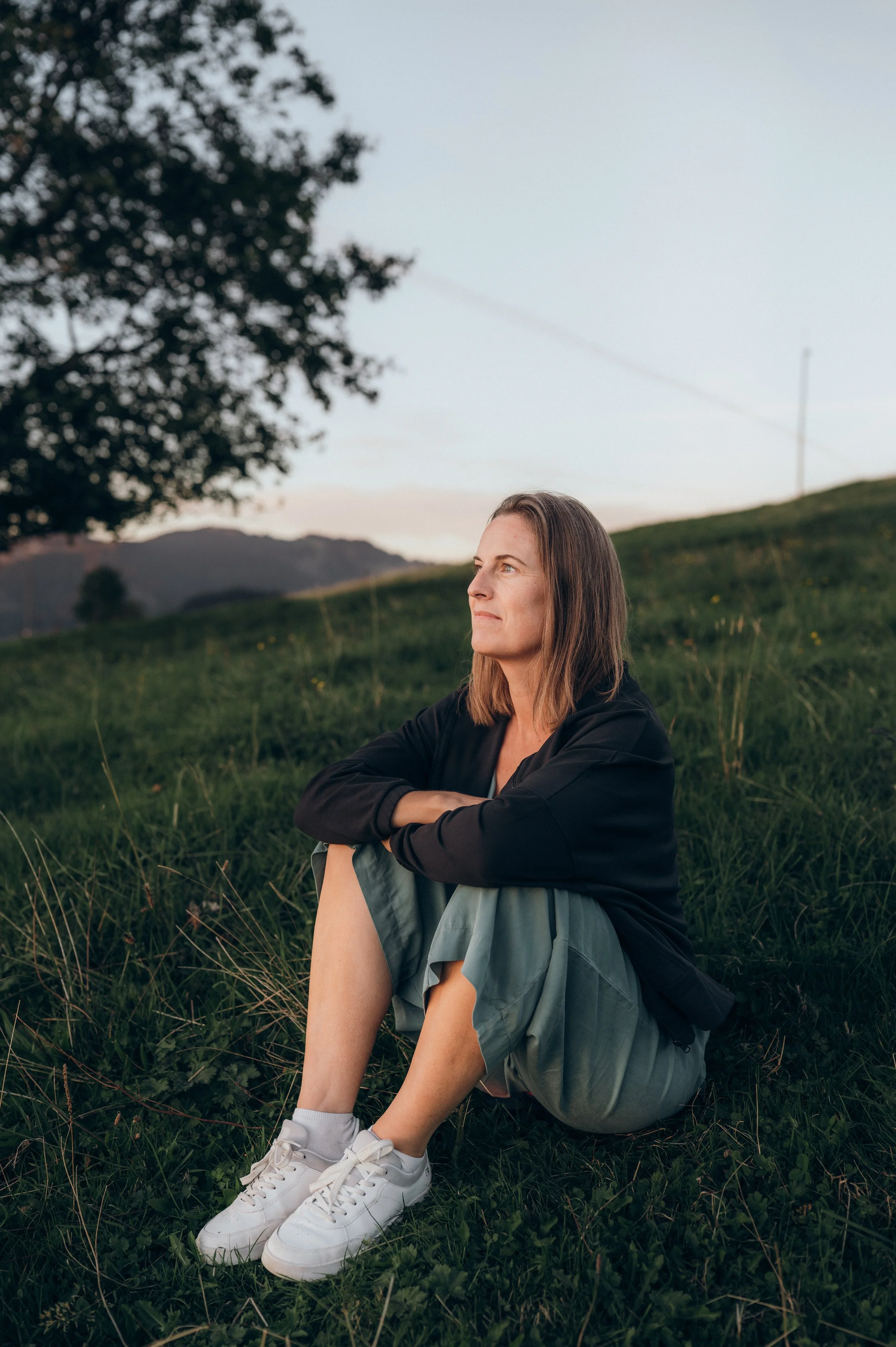 Junge Frau sitzt entspannt in einem grünen Gelände, mit Blick auf die Berge im Hintergrund, umgeben von Natur bei Sonnenuntergang.