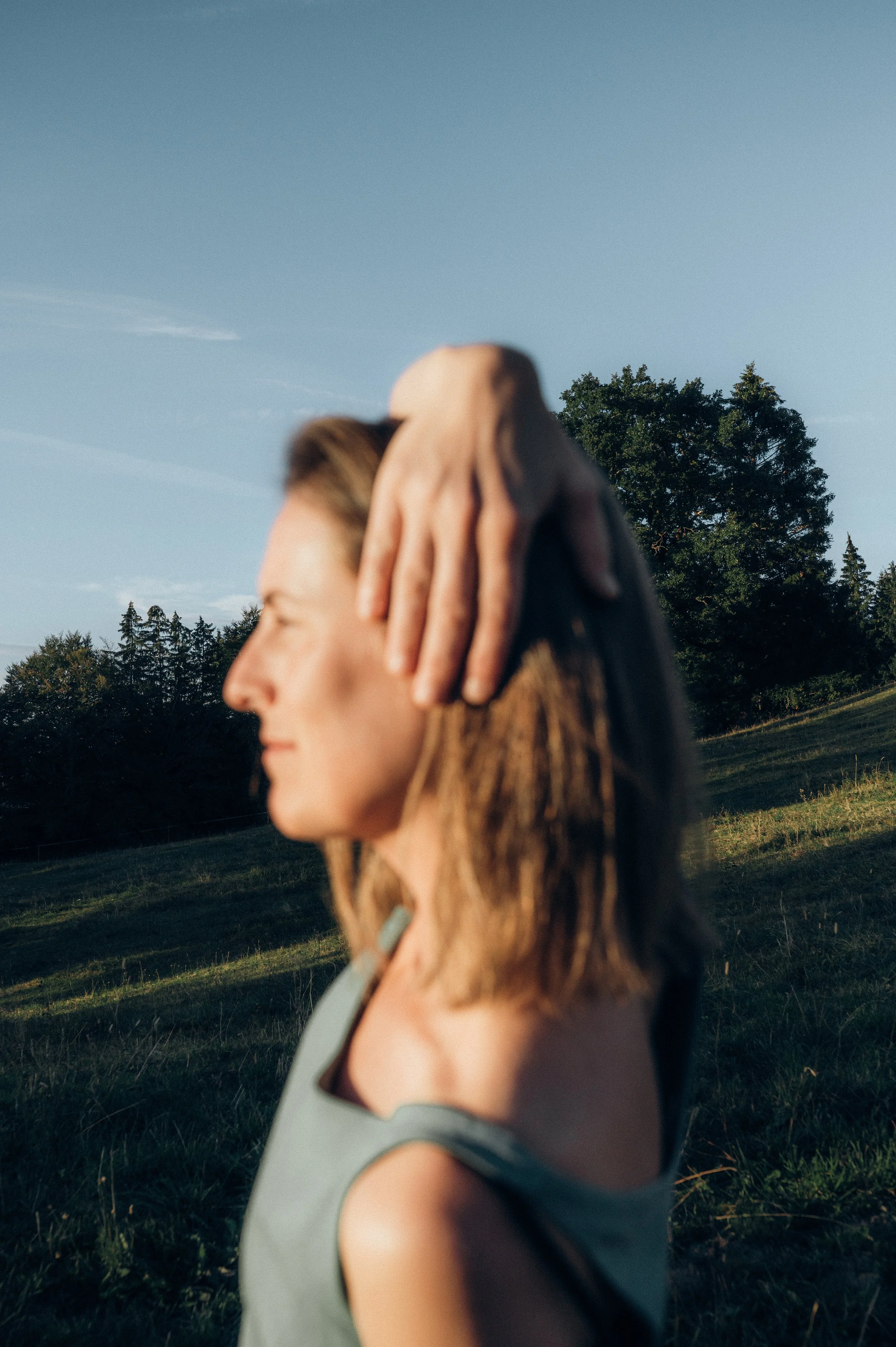 Porträt einer Frau mit braunen Haaren, die an einem sonnigen Tag im Freien steht, mit einer Hand auf ihrem Kopf, vor einer Landschaft mit Bäumen und blauem Himmel.